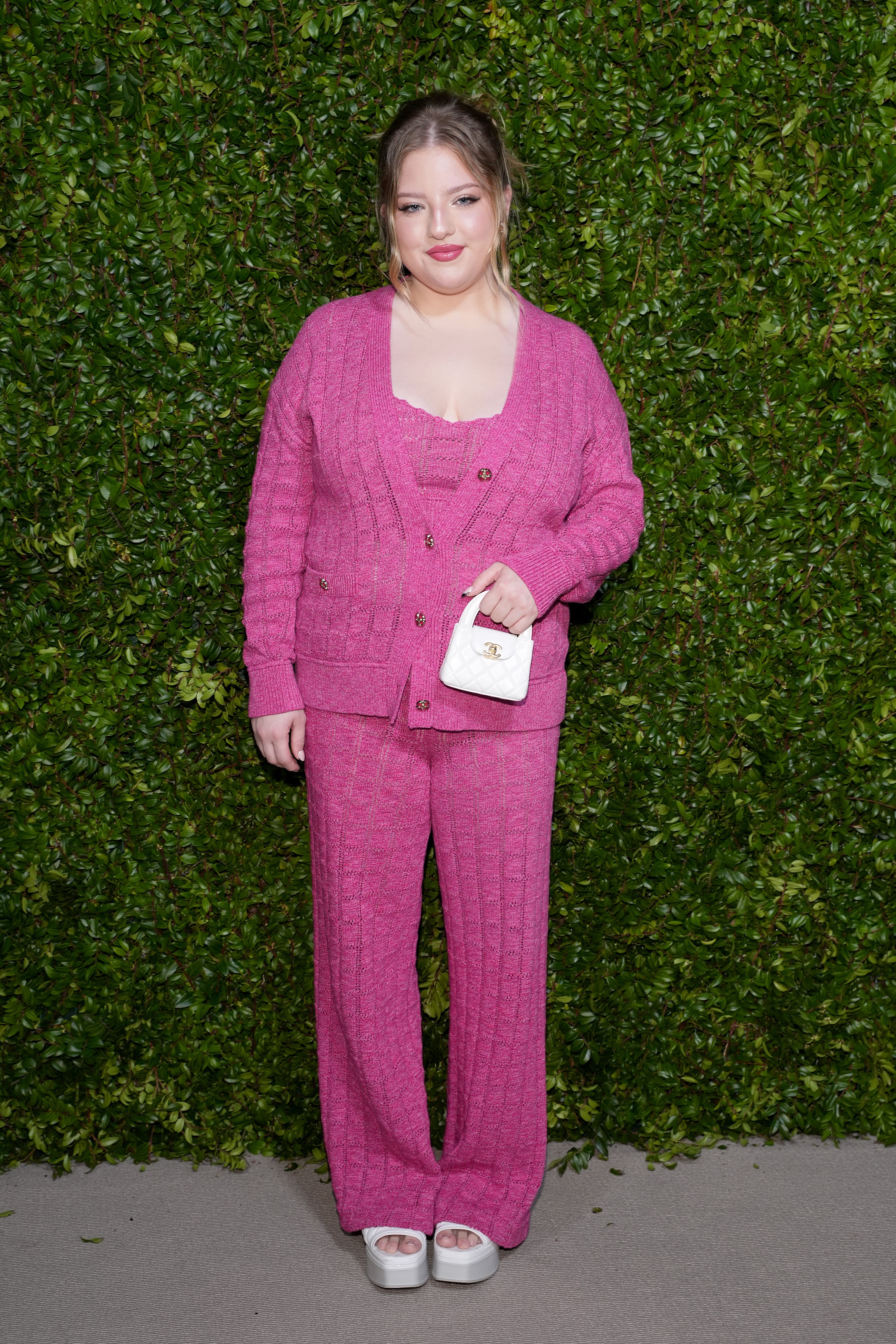 Person lasting  successful  beforehand   of a greenish  leafy wall, wearing a stylish matching knit set, featuring a cardigan and pants, holding a tiny  handbag