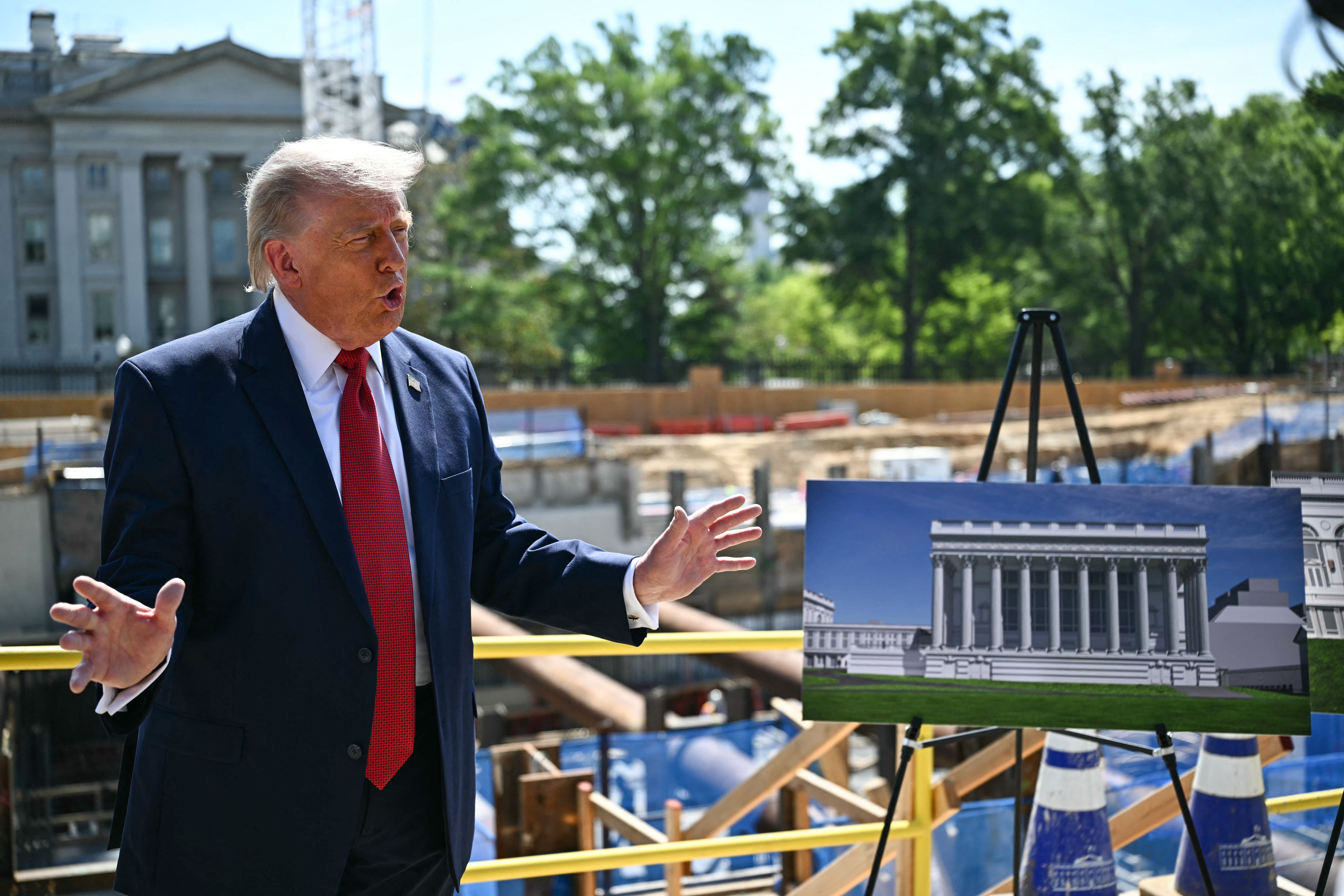 A man in a suit talks at a construction site with a rendering of a neoclassical building on display, suggesting future development plans