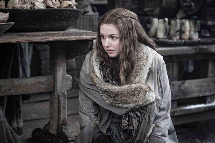 Woman in medieval-style fur-trimmed cloak sits in a rustic setting, looking to the side thoughtfully