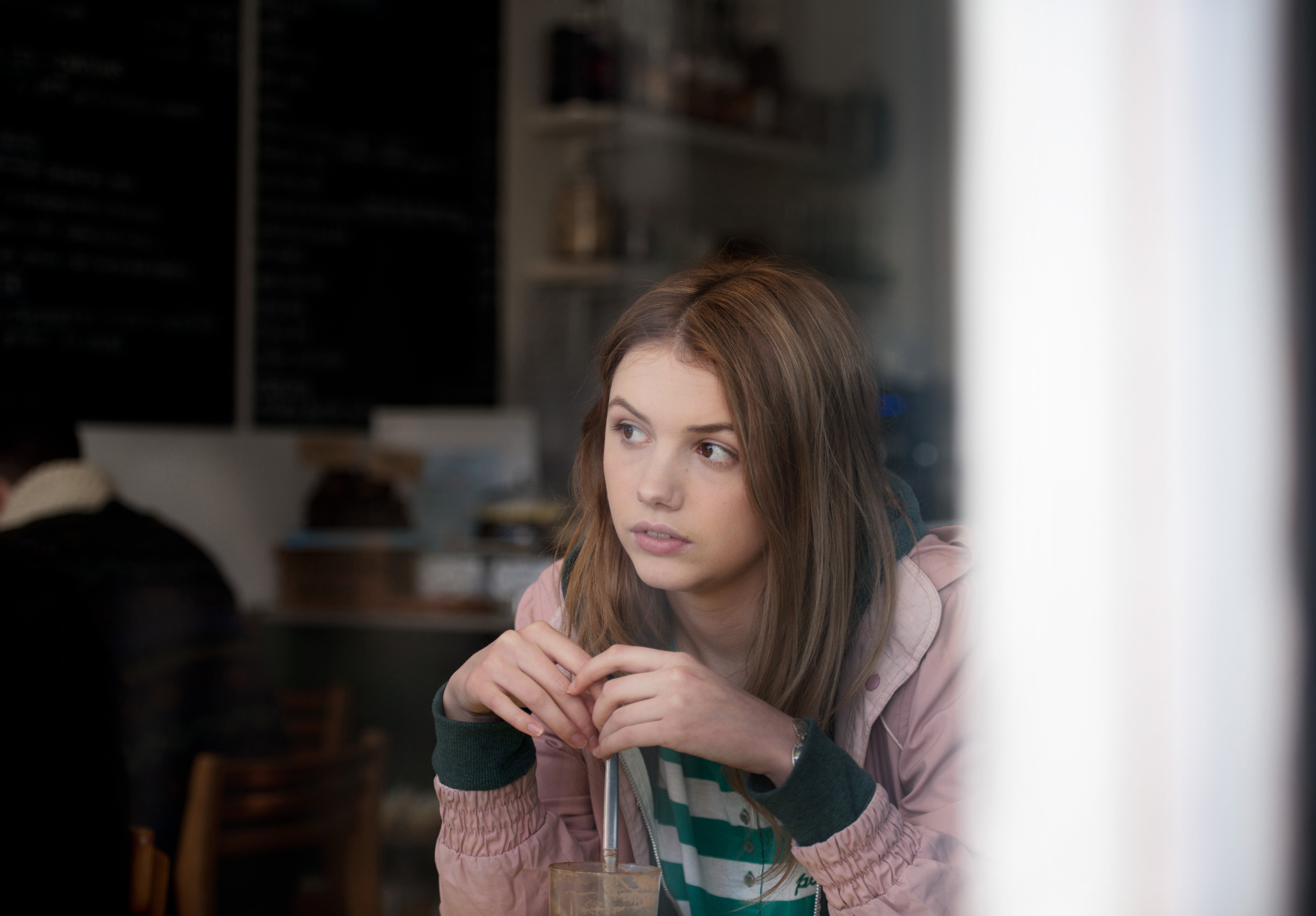 Person in a café, looking thoughtfully out of the window, wearing a casual striped shirt and a jacket