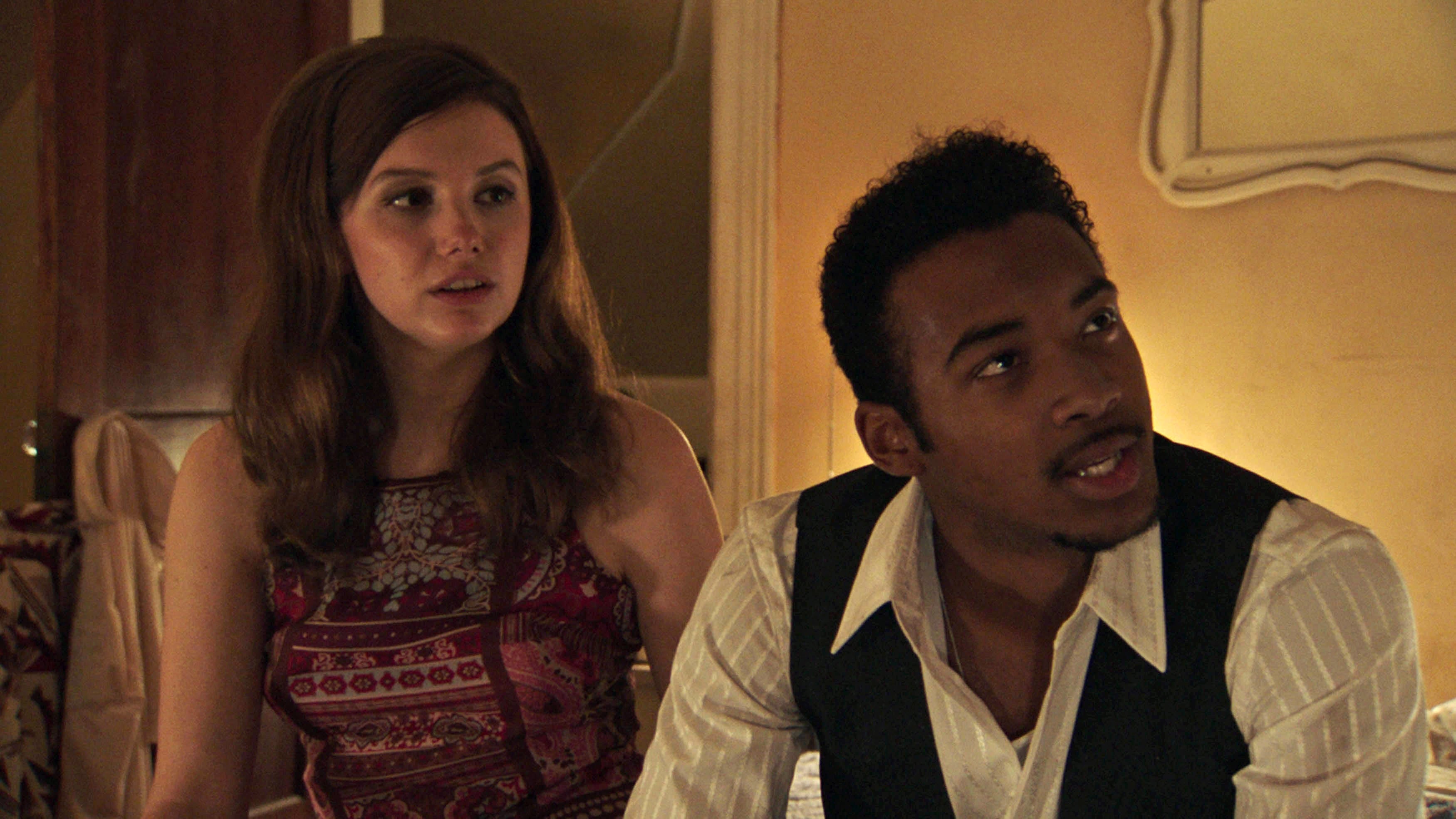 Two people are sitting together indoors, looking in the same direction, engaged in a conversation