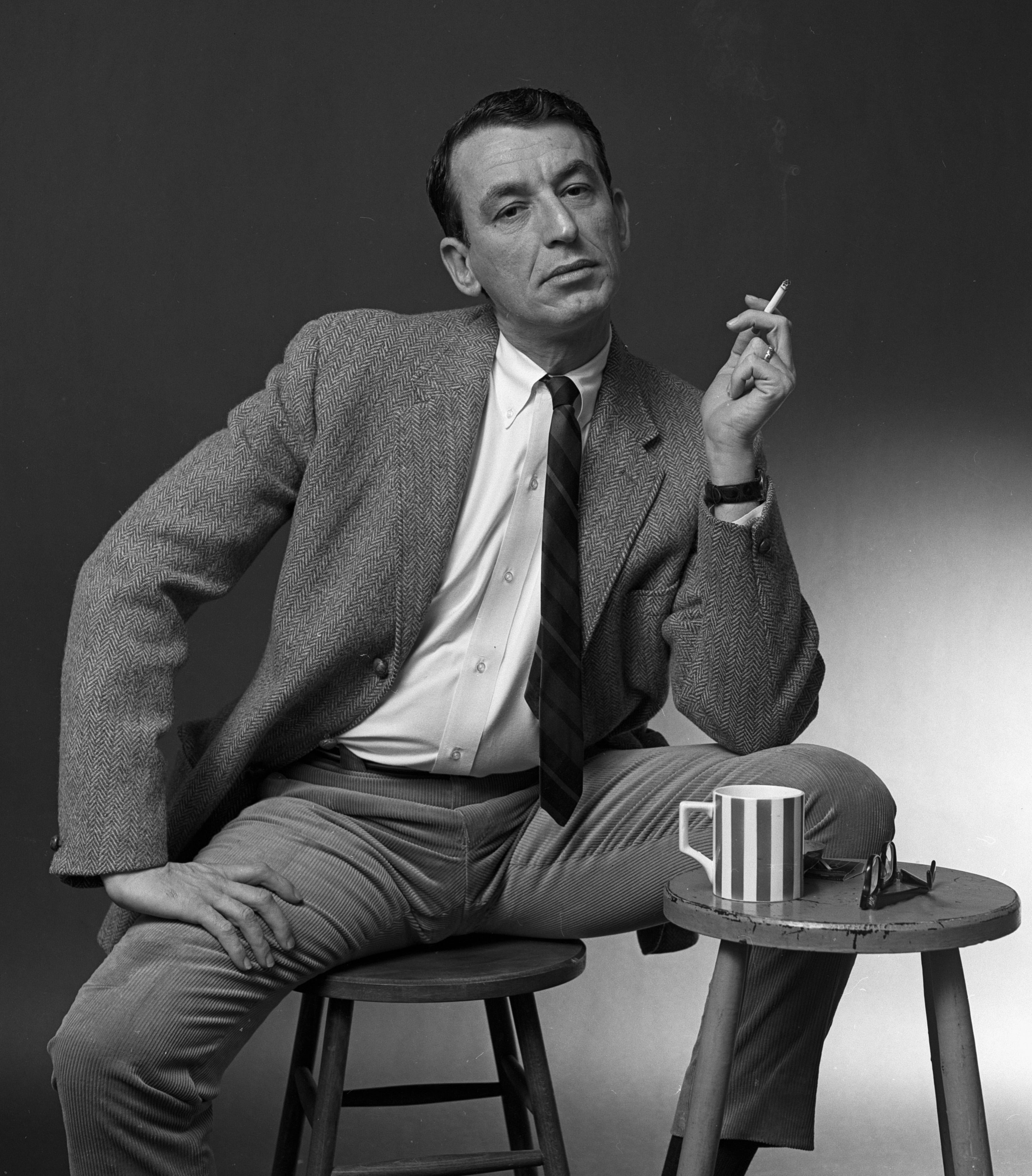 Person successful  a ceremonial  suit   sits connected  a stool, holding a cigarette, with a mug and tiny  array  nearby