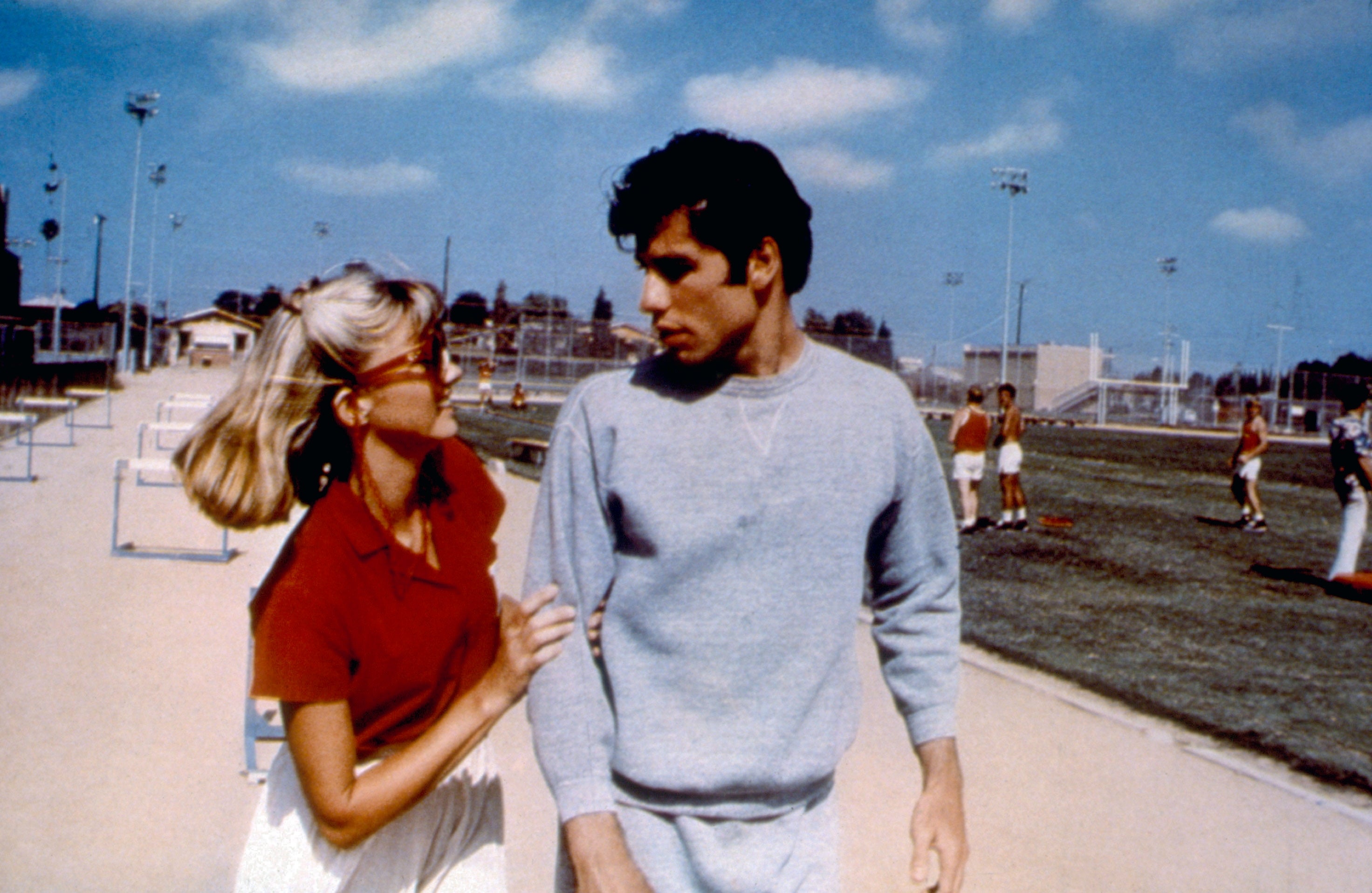Olivia Newton-John successful a reddish apical and skirt talks to John Travolta successful a grey sweatshirt, connected a sports tract with hurdles and radical successful the background