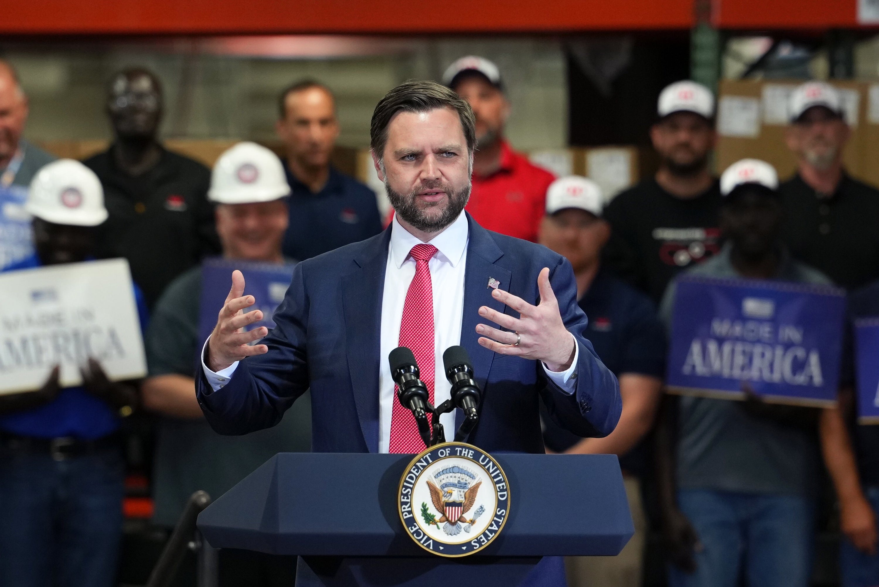A antheral   successful  a suit   and necktie  speaks astatine  a podium with microphones, surrounded by radical   holding "Made successful  America" signs