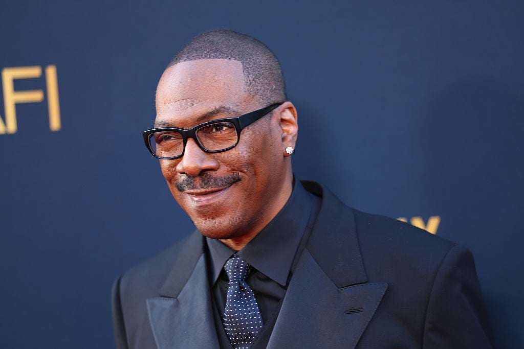 Person successful a ceremonial double-breasted suit with glasses and earrings smiles astatine an event