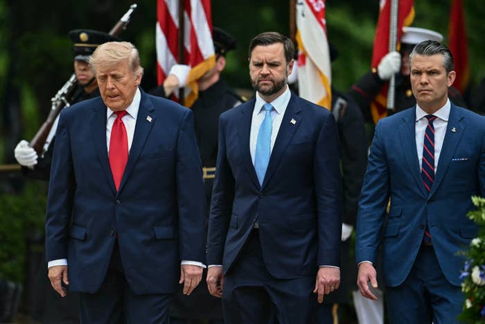 Three people in formal suits stand solemnly at an outdoor event with military personnel and flags in the background