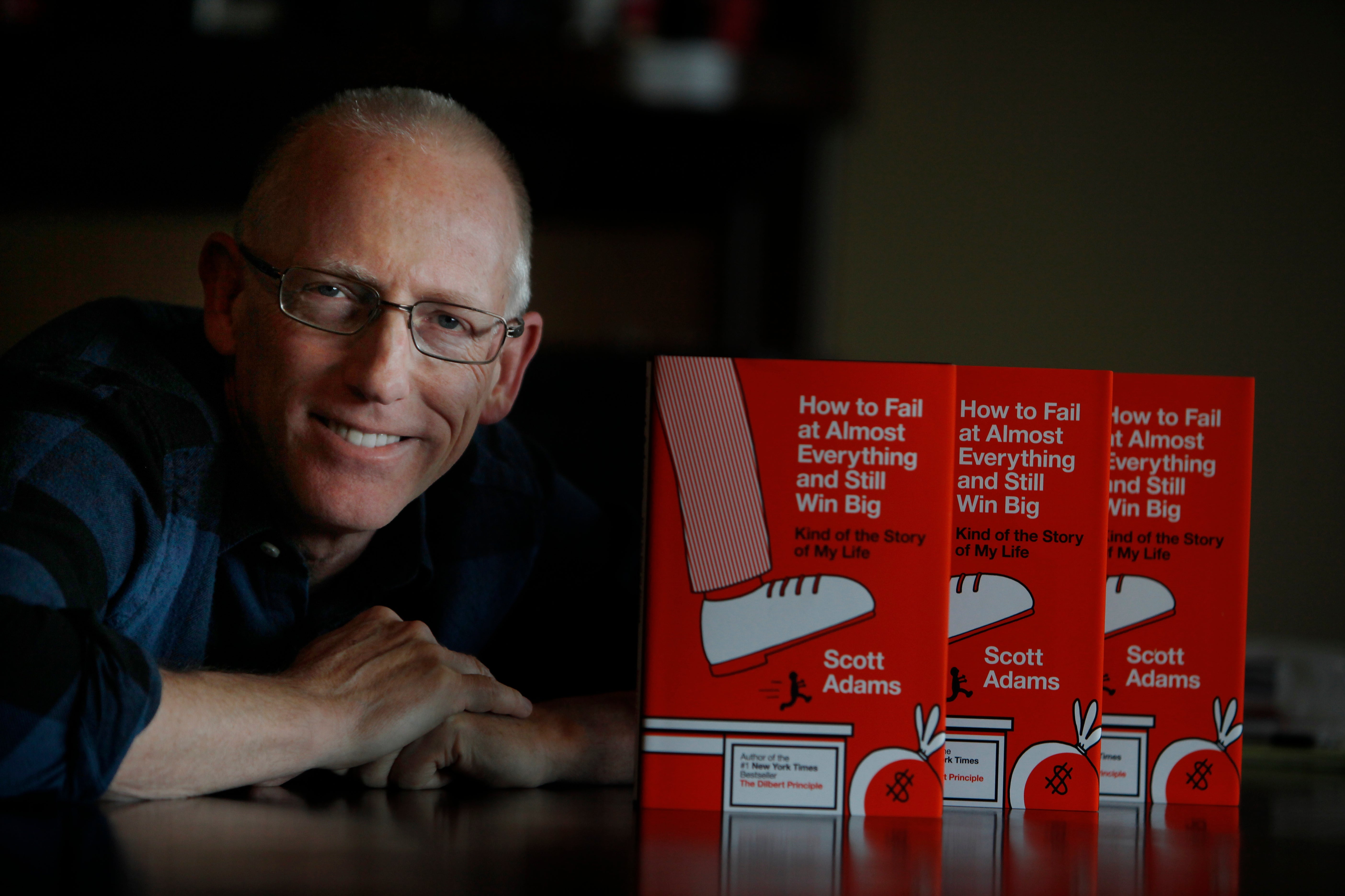 Dilbert creator smiling next to copies of a book titled "How to Fail at Almost Everything and Still Win Big" by Scott Adams