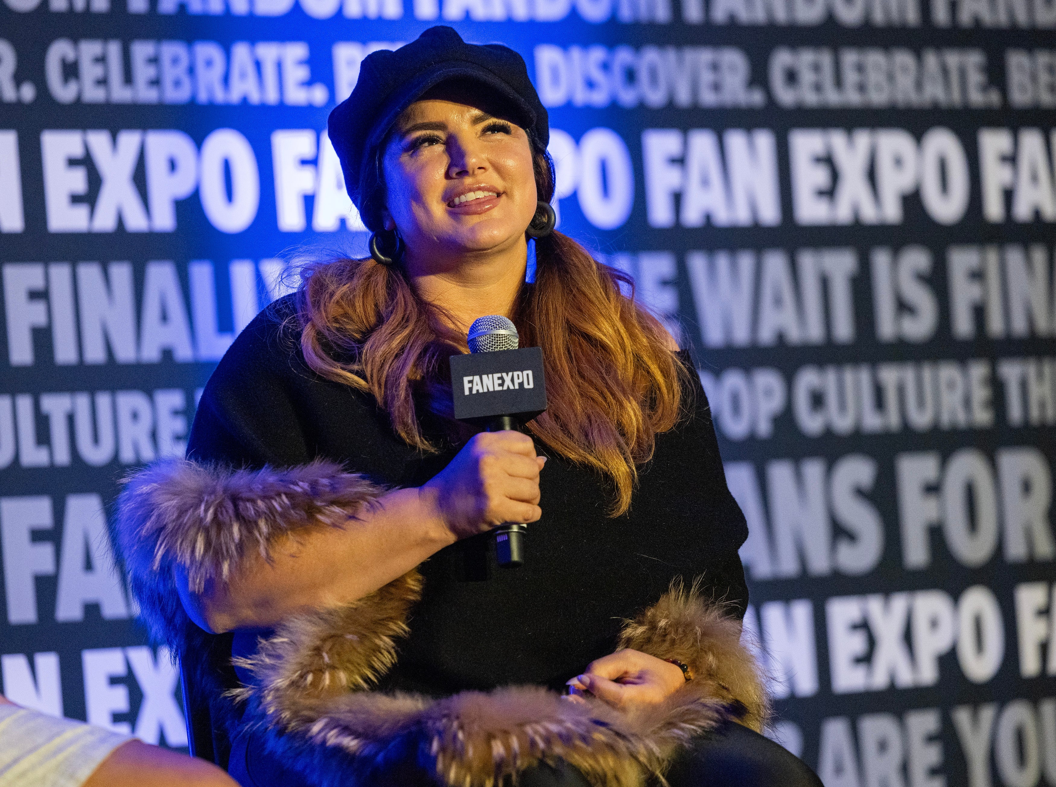 Gina Carno holding a microphone at a Fan Expo event, wearing a furry-trimmed outfit and a cap, seated in front of a backdrop with repeated text
