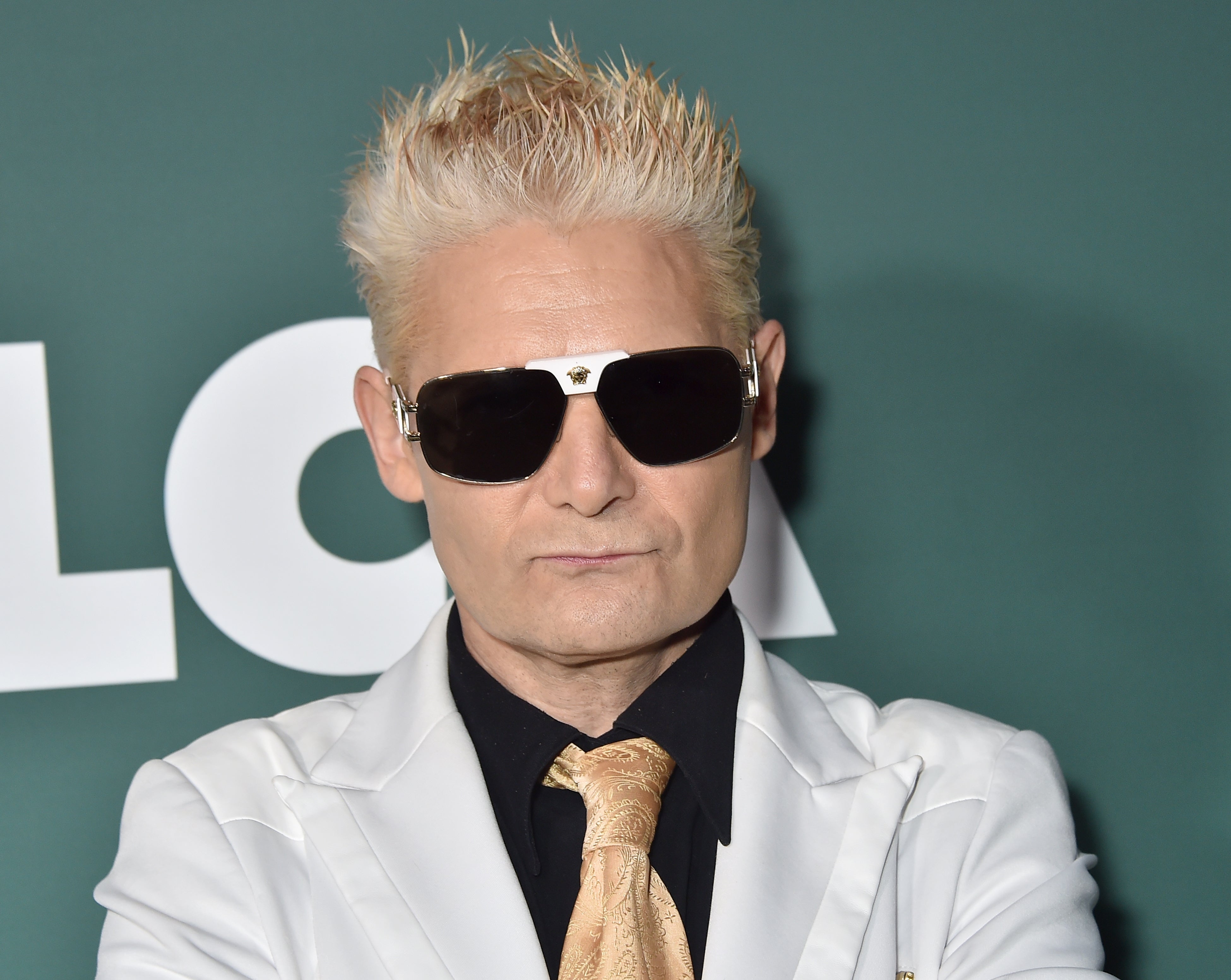 Corey Feldman wearing sunglasses and a white suit with a gold tie, standing against a backdrop