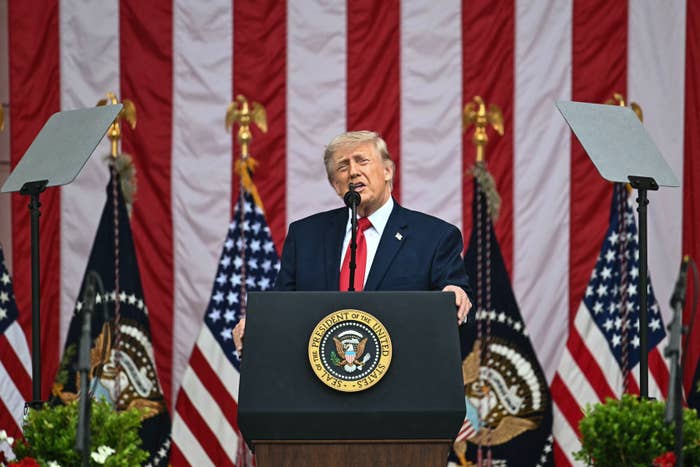 Person speaking at a podium with presidential seal, flanked by U.S. flags and large red and white striped backdrop