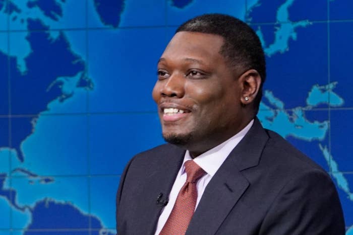 Two TV hosts in suits sit at a news desk, smiling. A world map is displayed in the background