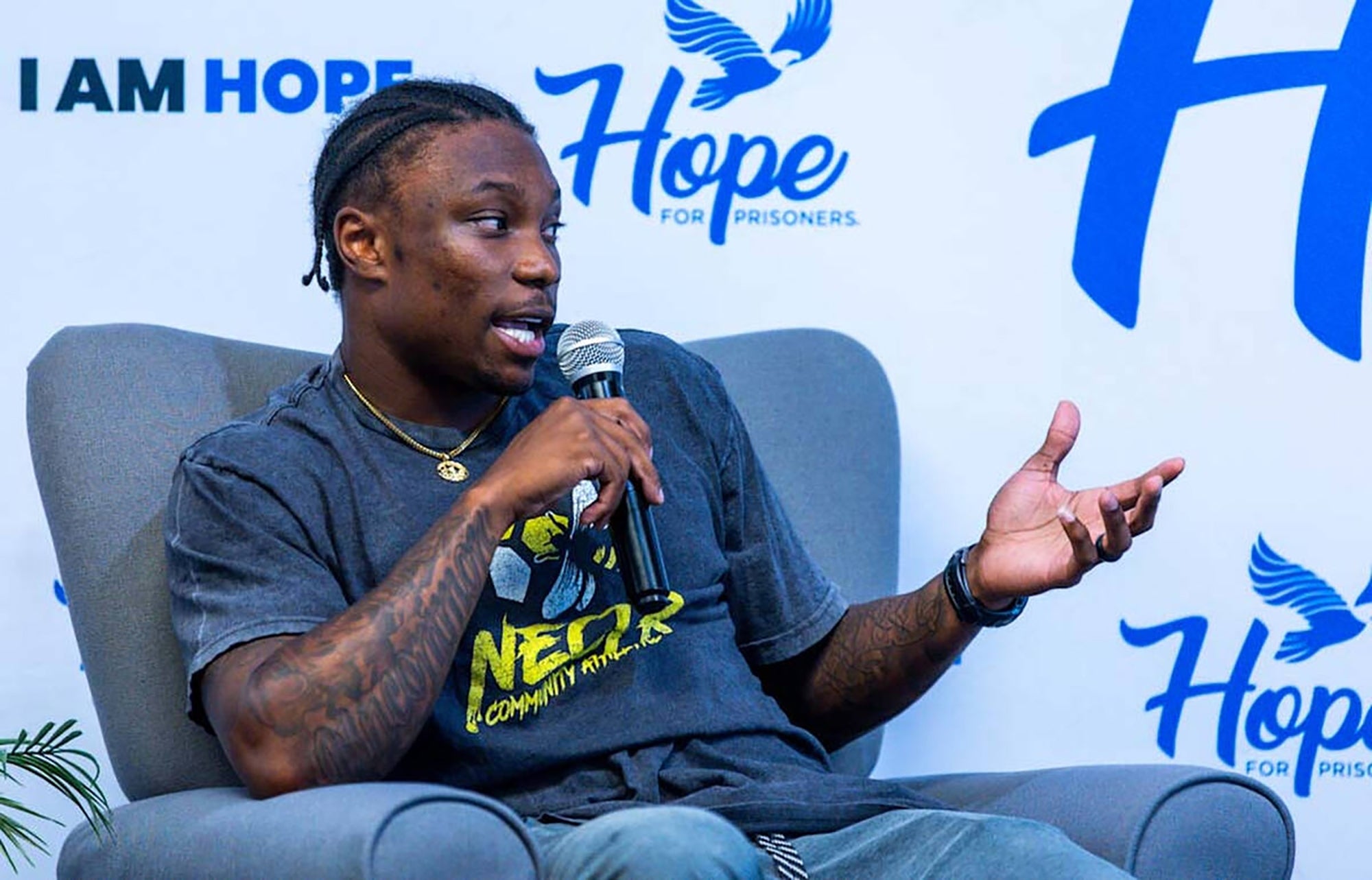 Person speaking on stage, wearing a graphic t-shirt, holding a microphone. Background features logos and text related to "Hope for Prisoners."