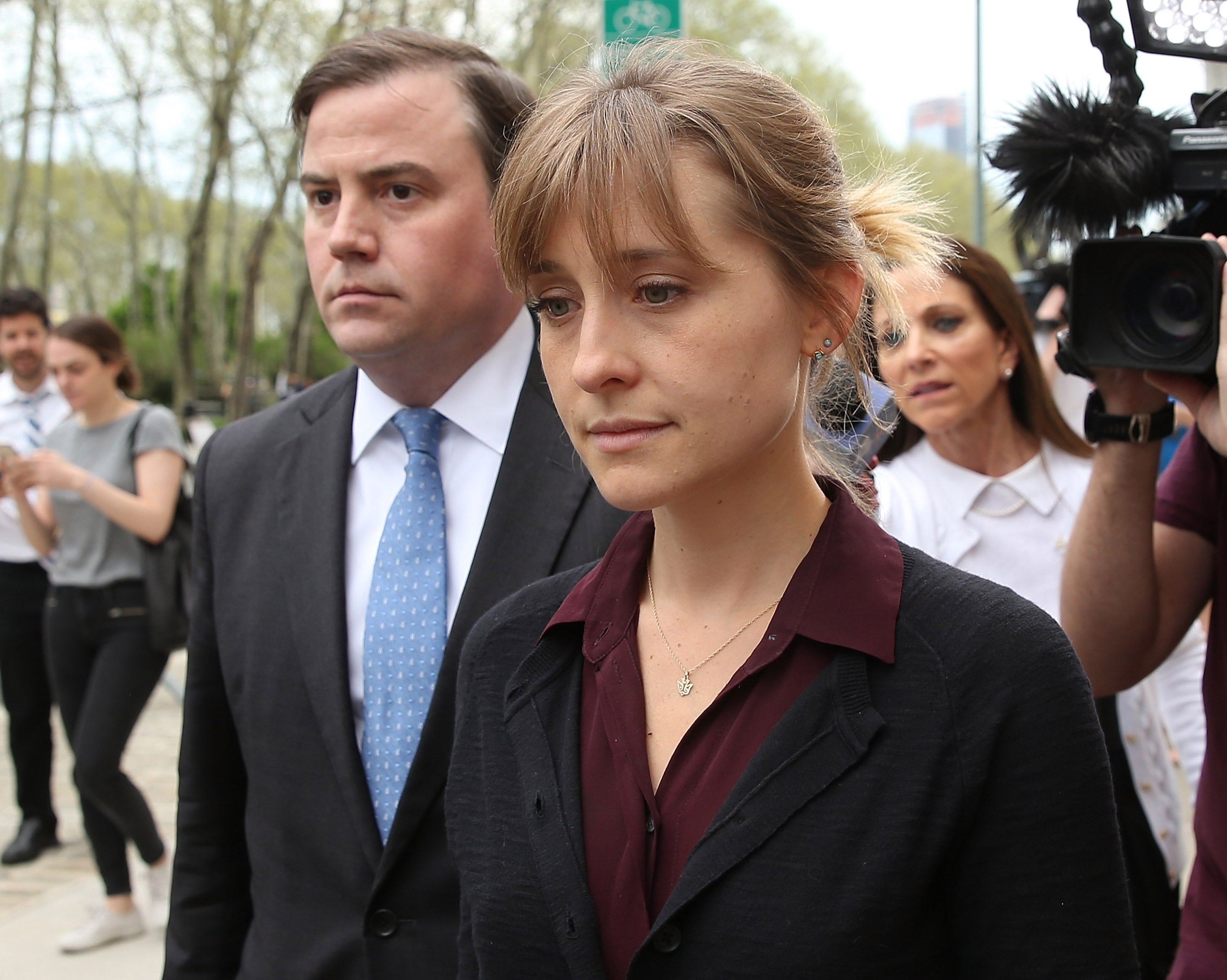 Allison Mack in professional attire walks with a group outside, appearing serious. People and cameras surround her