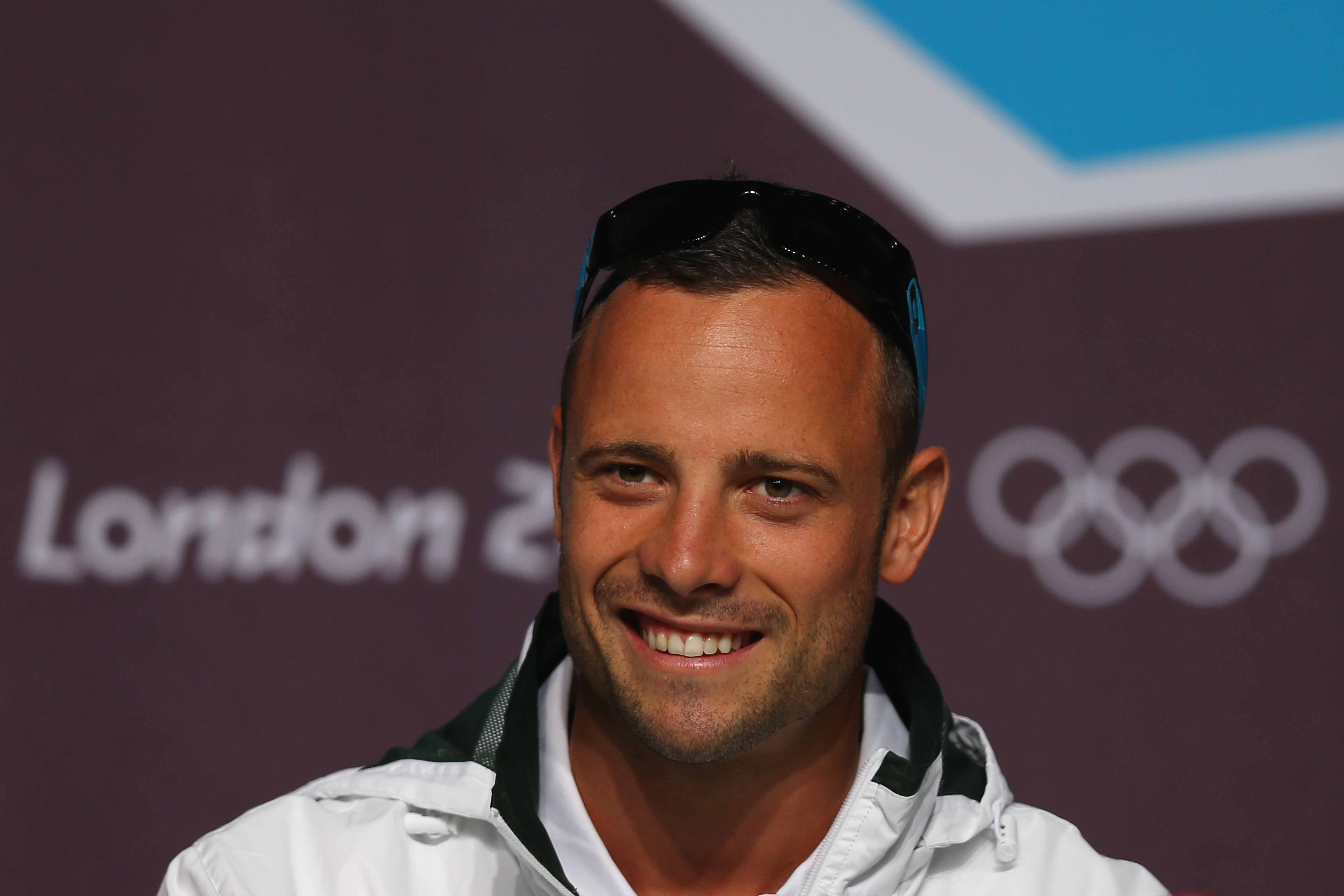 Oscar Pisotorius smiles at a press event with an Olympic backdrop. He's wearing a jacket and sunglasses on his head
