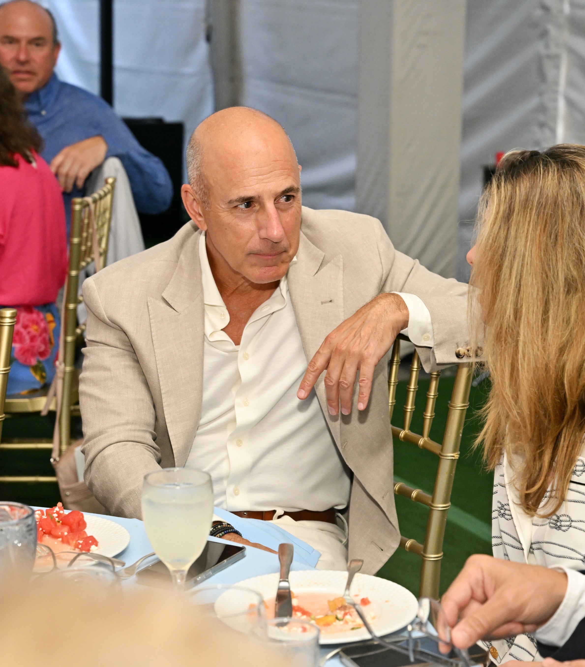 Matt lauer  in a beige suit sits at a dinner table, conversing with a woman at a social event