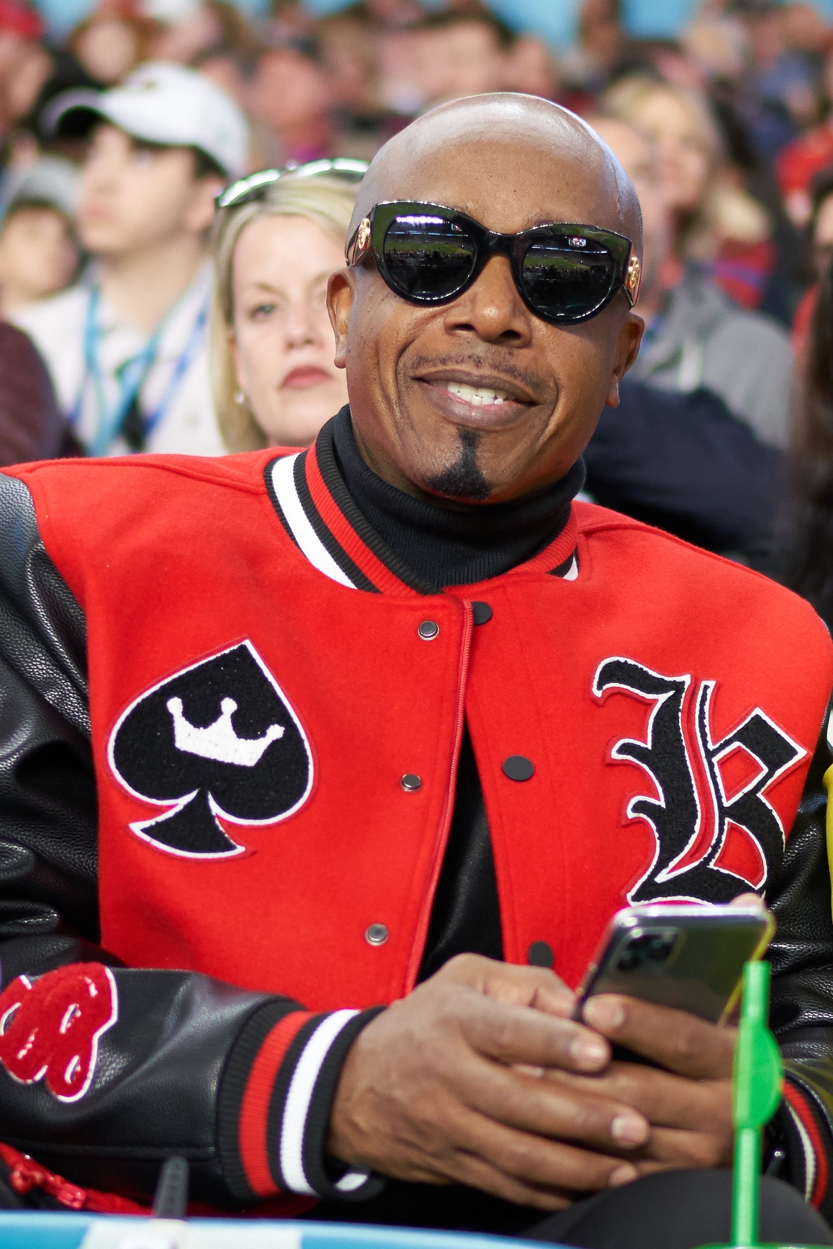 MC Hammer at an event, wearing sunglasses and a red varsity jacket with card suit designs, holding a phone