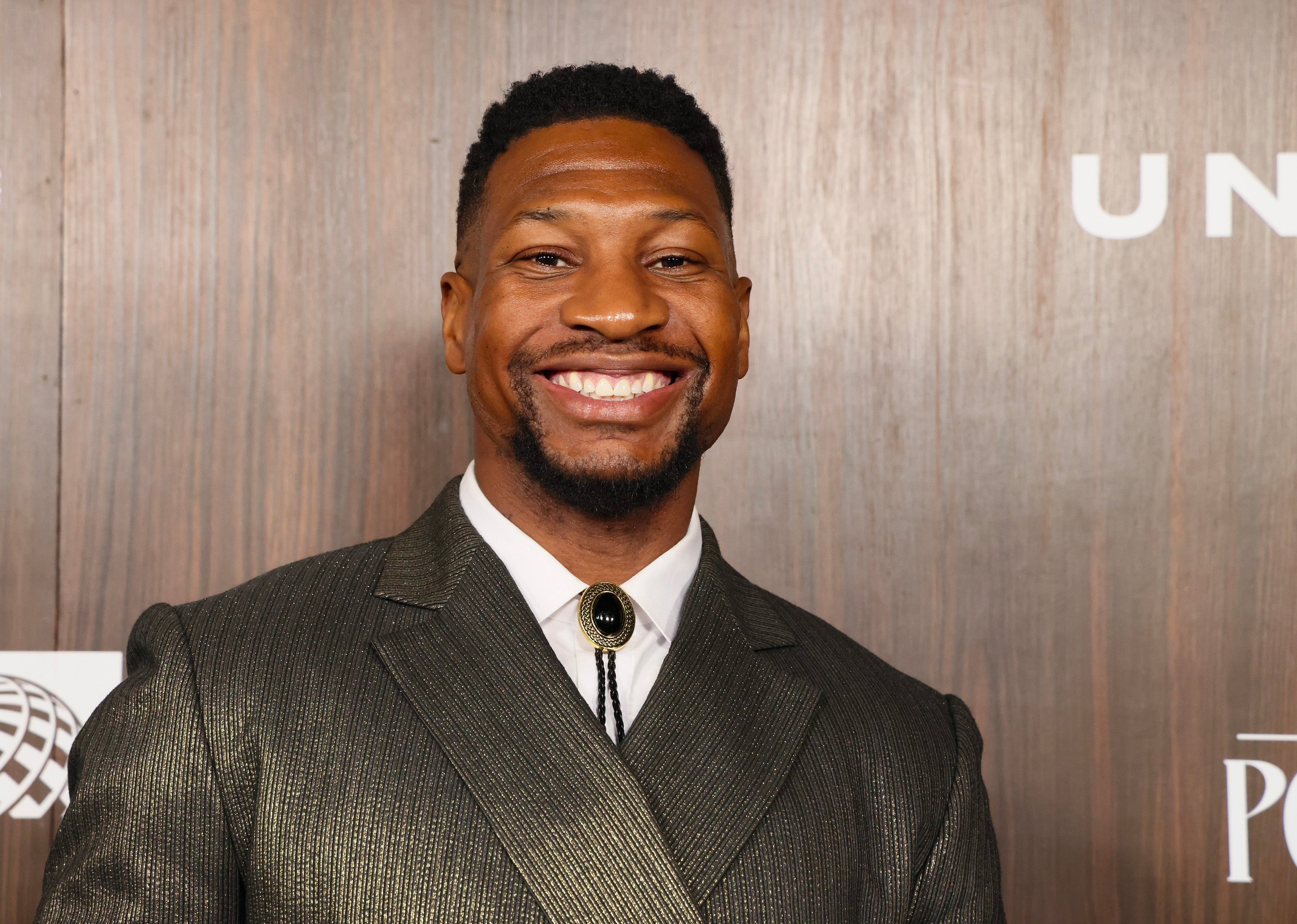 Jonathan Majors in a shiny suit smiles at an event with wooden backdrop