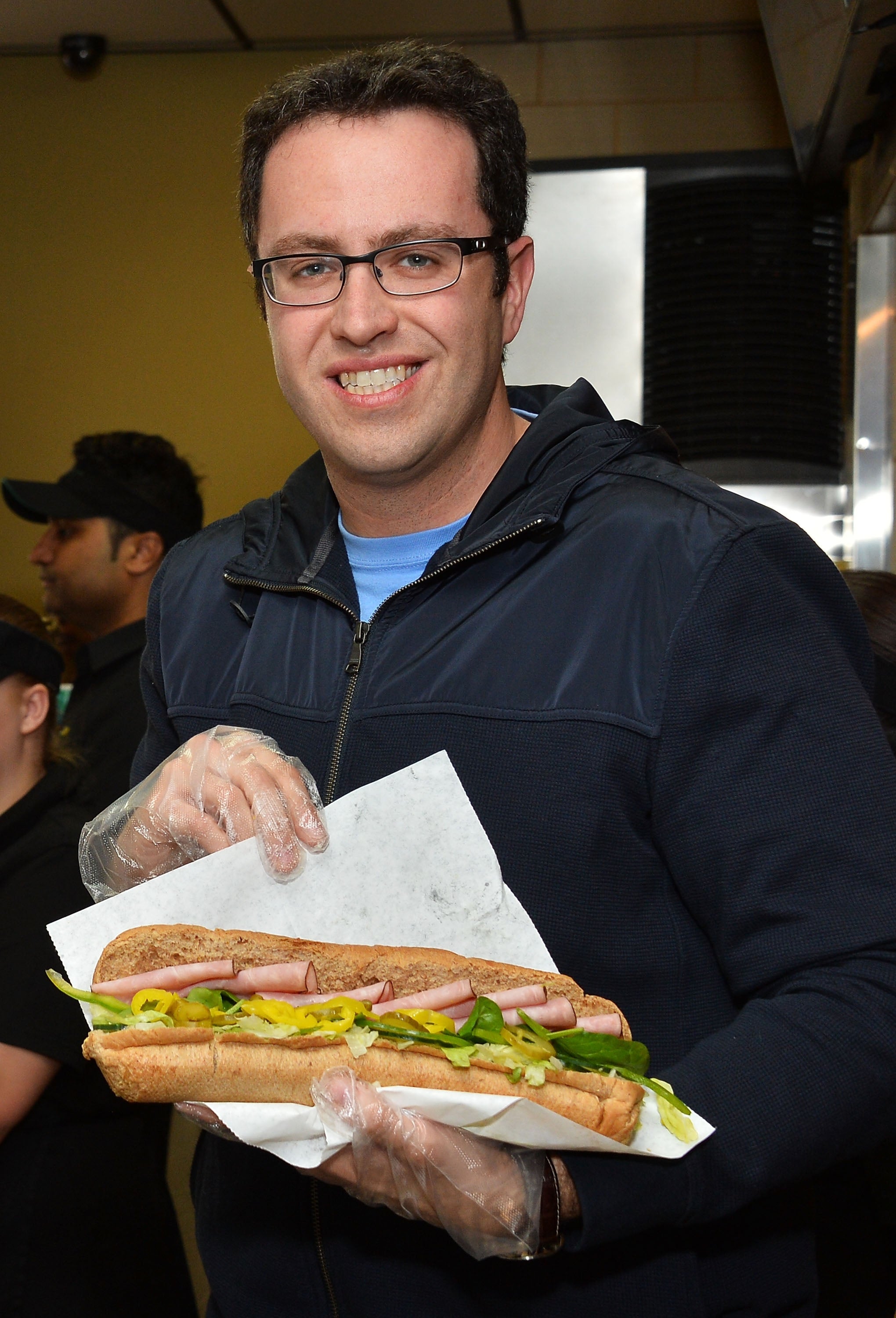 Jared of Subway fame holds a large sub sandwich, wearing clear gloves and a casual jacket. Other people are visible in the background