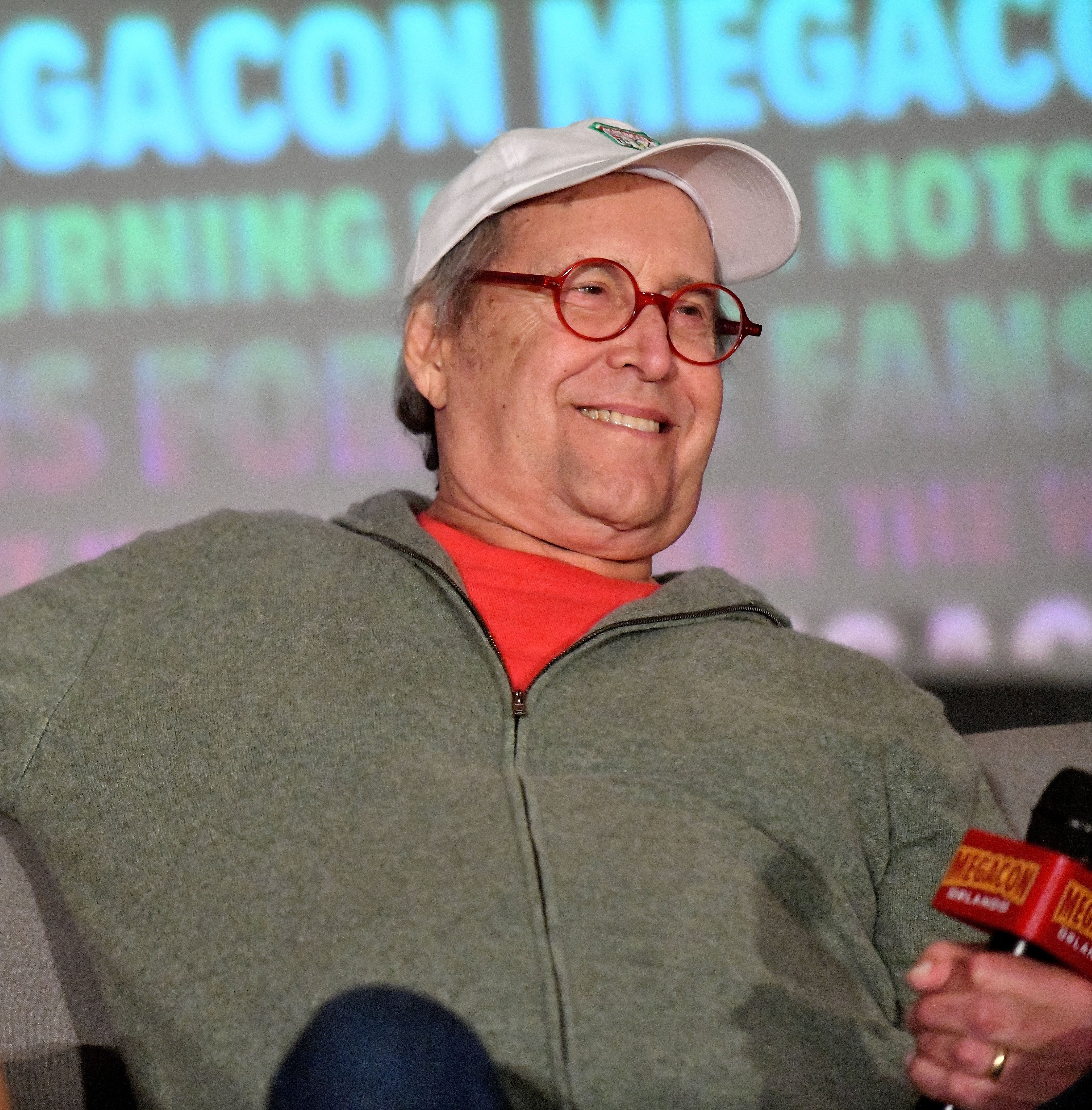 Chevy Chase wearing a cap and glasses, sitting with microphones, in front of an event backdrop