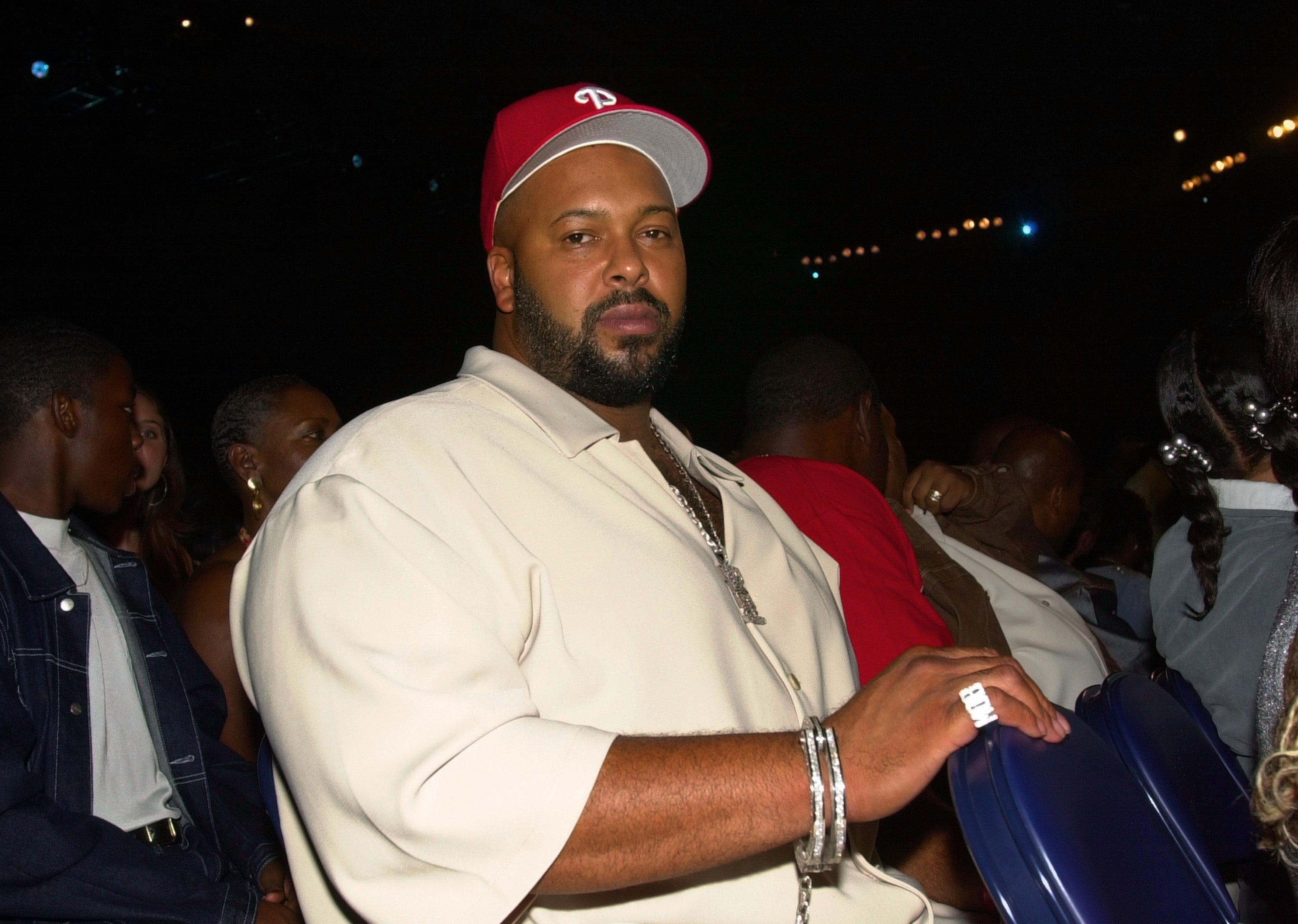 Suge Knight wearing a red cap and casual attire, sitting in a crowded event with others around him