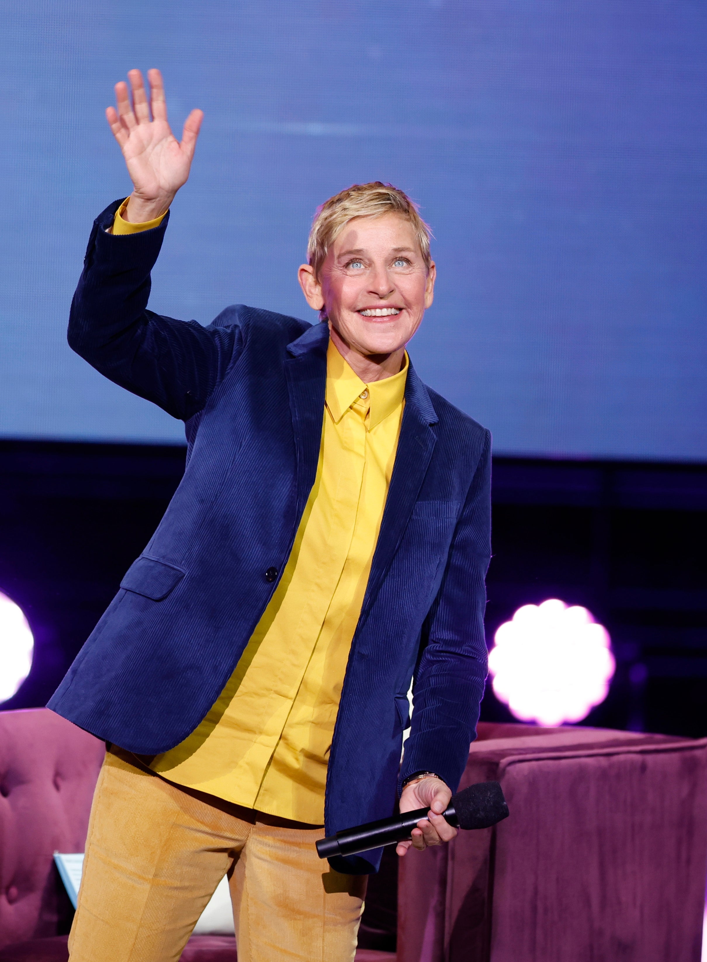 Ellen Degeneres in a yellow shirt and blue blazer waves while holding a microphone, standing on stage with a purple chair in the background