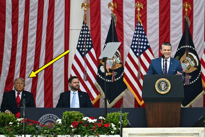 Man at podium speaks during an event with U.S. flags and seal; two seated guests listen. Formal setting indicates a significant event