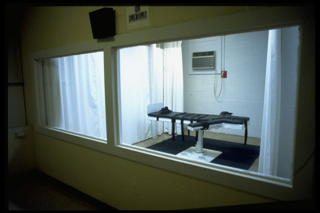Execution enclosure  with a viewing window, featuring a gurney with restraints. The country   has aesculapian  instrumentality  and is viewed from an adjacent room