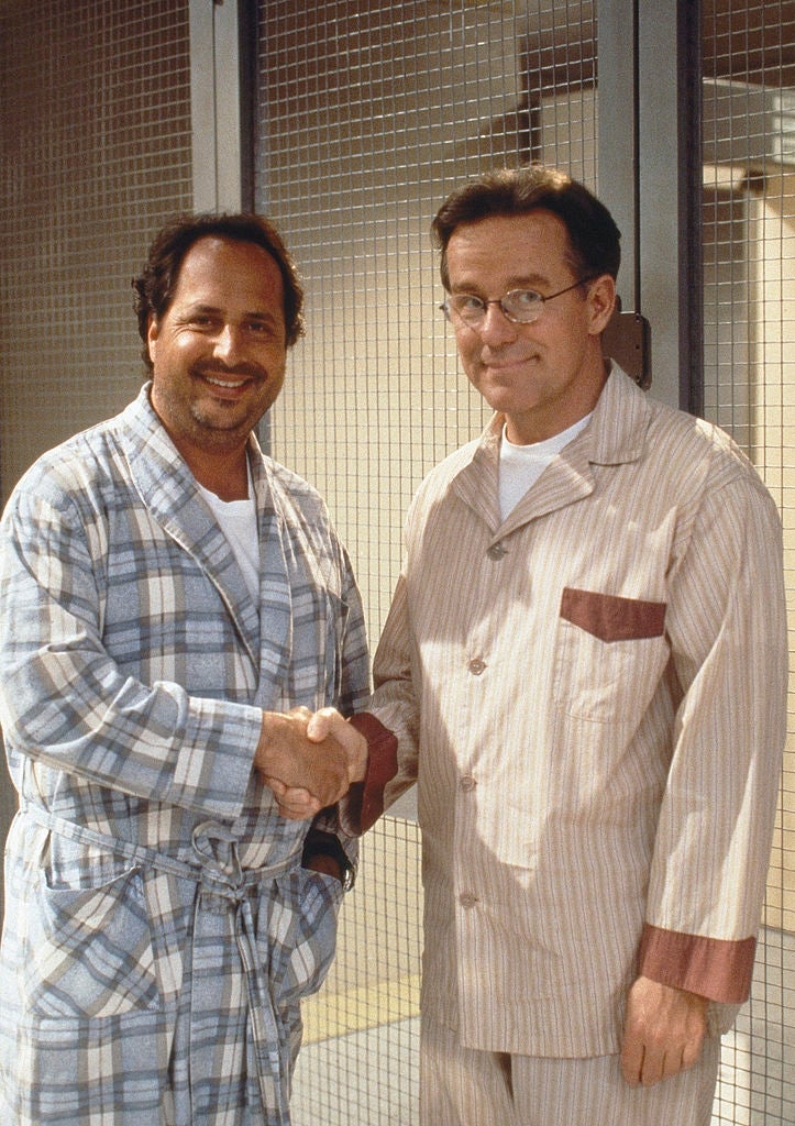 Two men successful  pajamas shingle  hands successful  a checkered room. One wears a plaid robe, the different   successful  striped pajamas with glasses. Both are smiling
