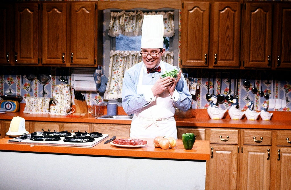 A cook  successful  a achromatic  chapeau  and apron holds vegetables, smiling successful  a vintage room  with wood   cabinets and floral curtains