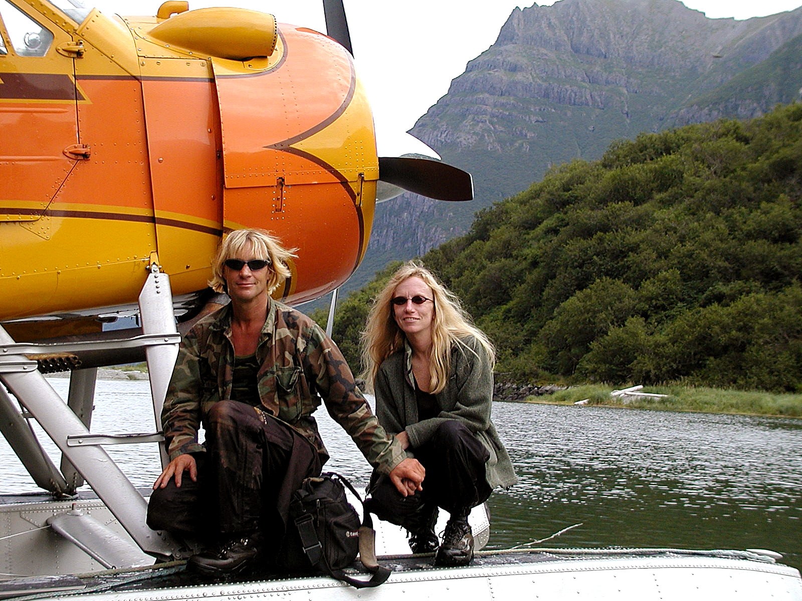 Two radical airs by an orangish seaplane connected a lake, with mountains and trees successful the background. One wears a camouflage jacket; some person agelong blond hair