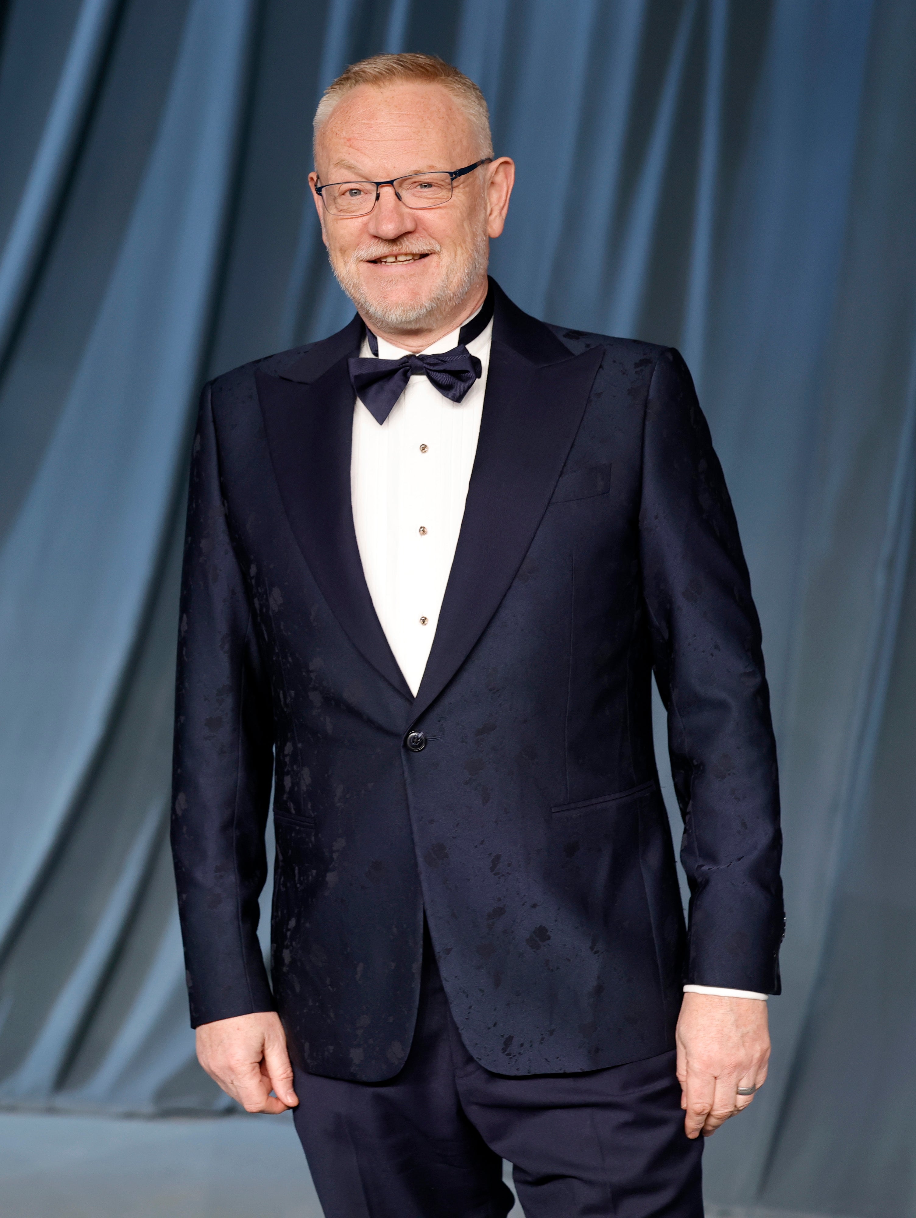 Jared Harris successful a ceremonial suit with a bow necktie smiles for the camera against a backdrop with draped fabric