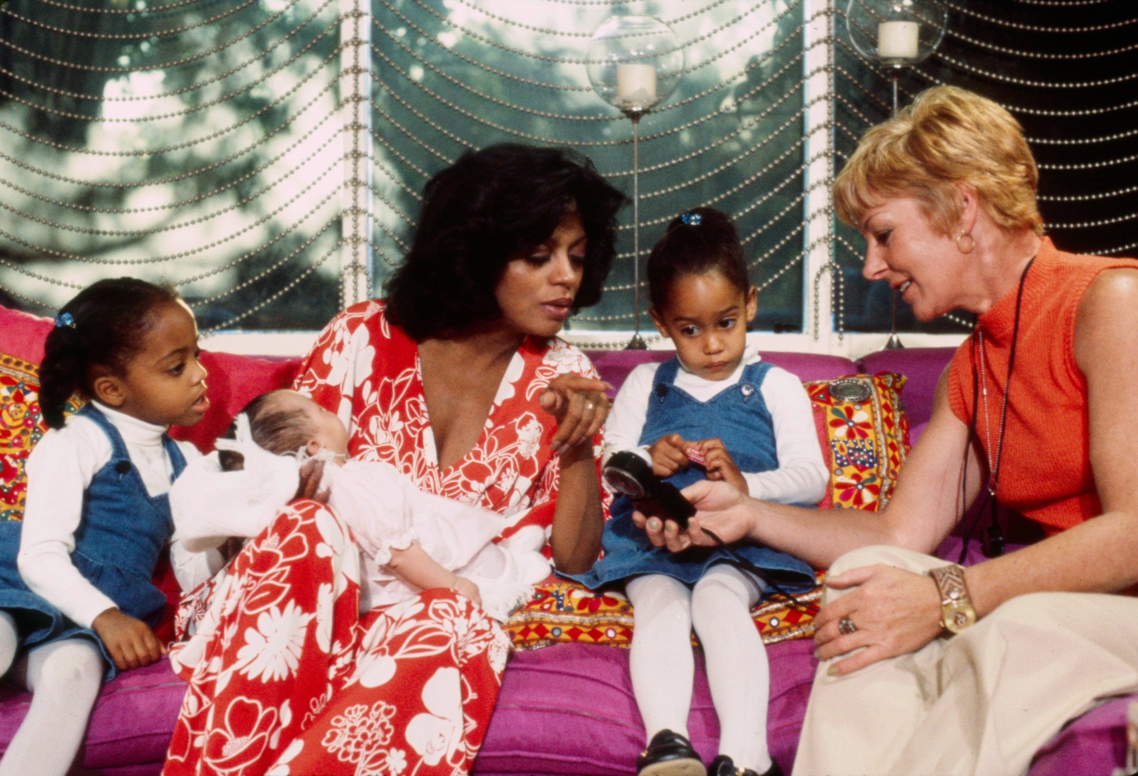 Diana Ross successful a floral formal sits with her 3 children and different pistillate connected a sofa.