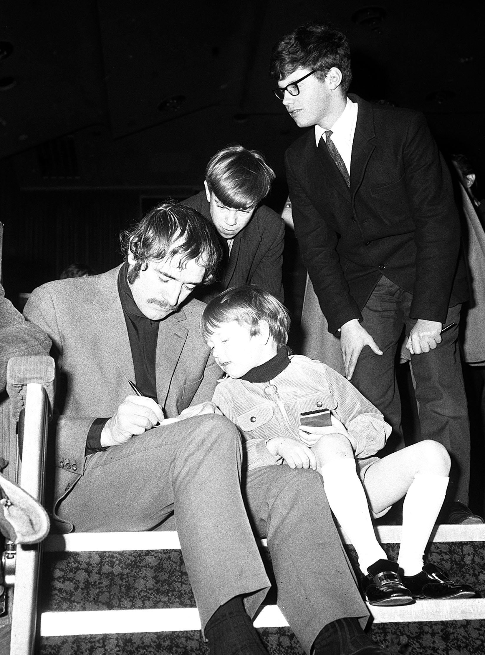 Richard Harris signs an autograph for a child, with 2 different children and a antheral lasting nearby