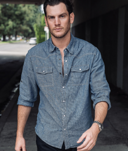 Person walking outdoors, wearing a casual denim button-up shirt with sleeves rolled up and dark pants. Urban backdrop visible