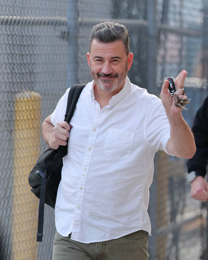 Man successful  casual attire, achromatic  shirt, holding keys and a container  implicit    shoulder, smiling and waving portion    walking outside