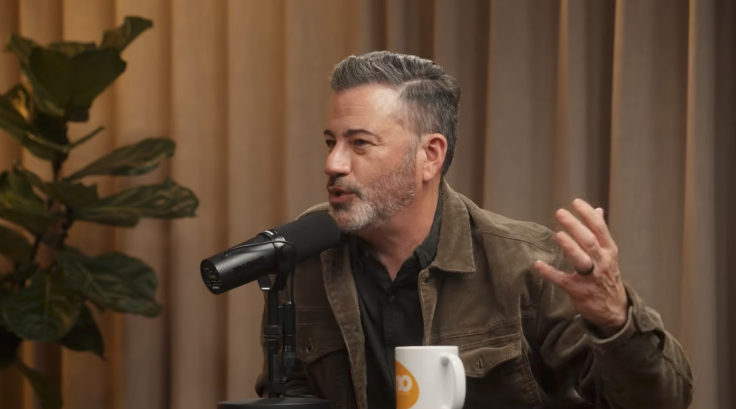 A idiosyncratic   speaks into a microphone successful  a podcast setting, gesturing with 1  hand. A mug is connected  the table