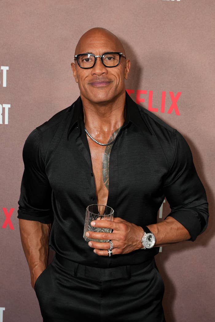 A man in a tailored black shirt and glasses, holding a drink on a red carpet, at a media event