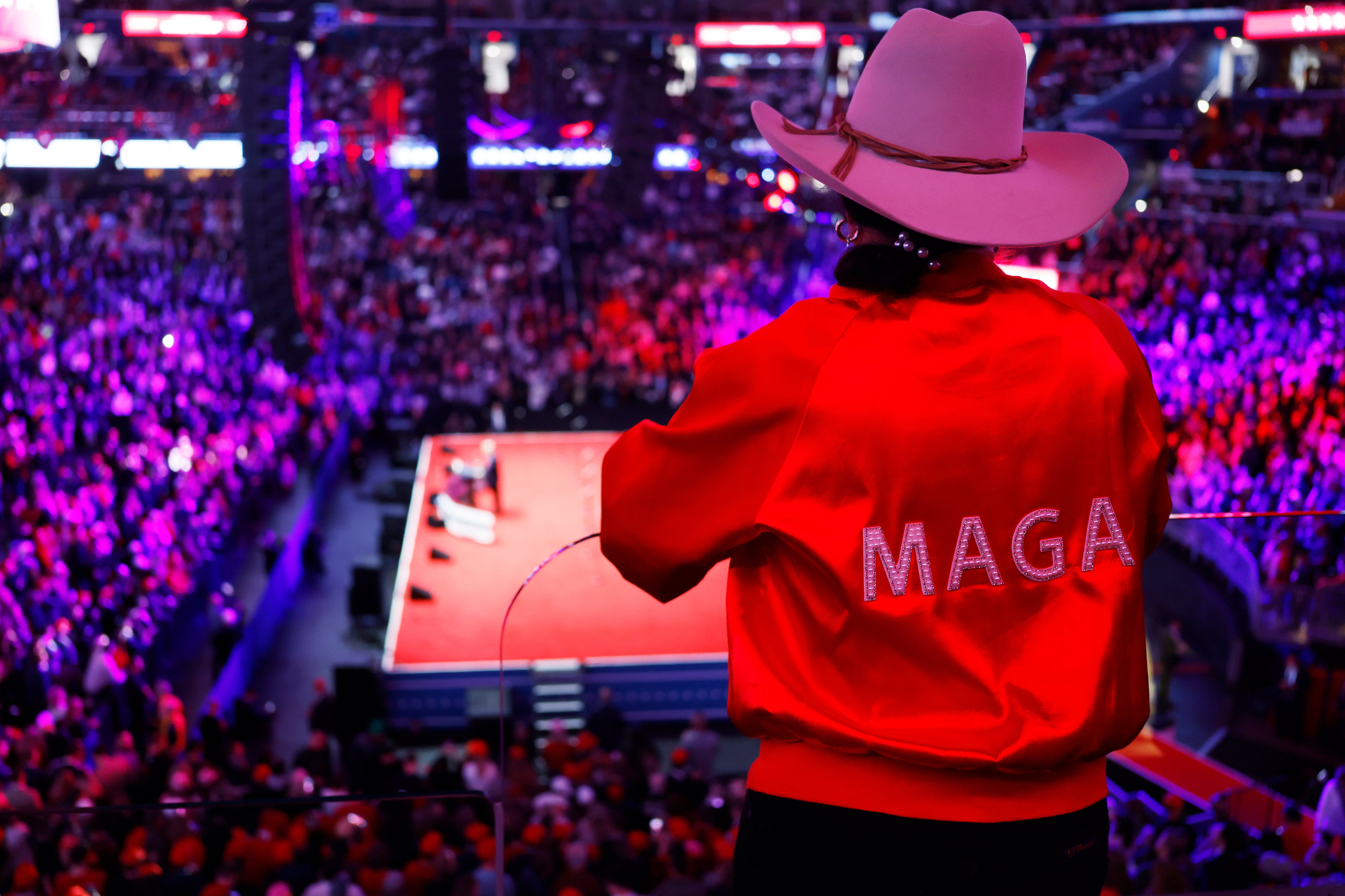 Person i cowboyhatt og MAGA-jakke har utsikt over en fullsatt arena med scene