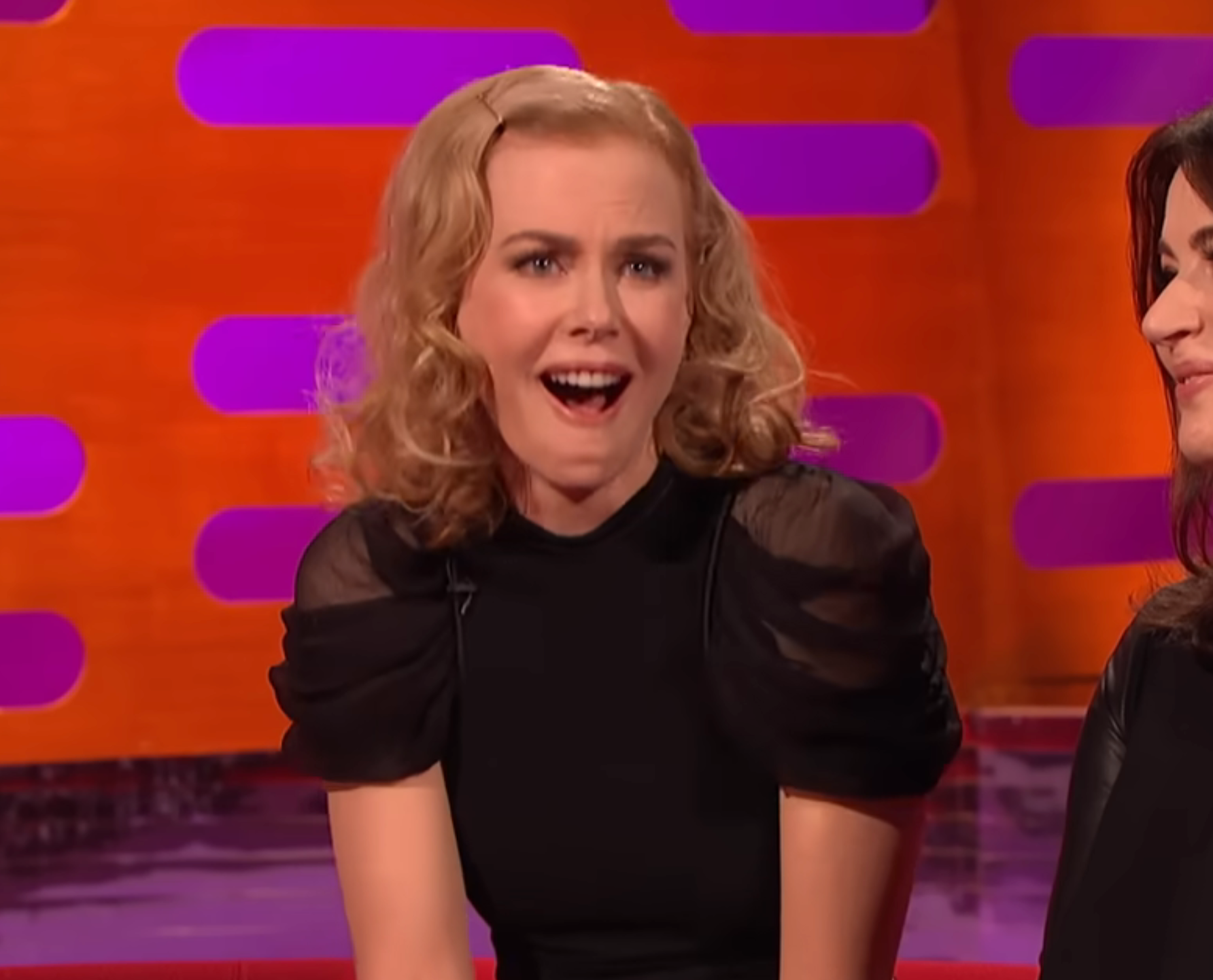 A person with wavy hair laughs animatedly while seated, wearing a stylish black dress with puffed sleeves