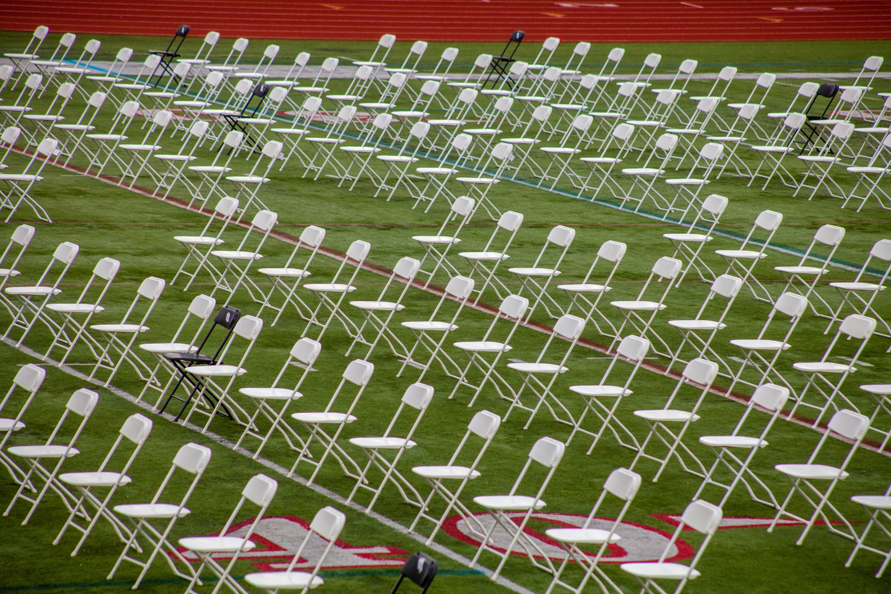 Rows of bare achromatic folding chairs are arranged connected a sports field, with a fewer achromatic chairs interspersed