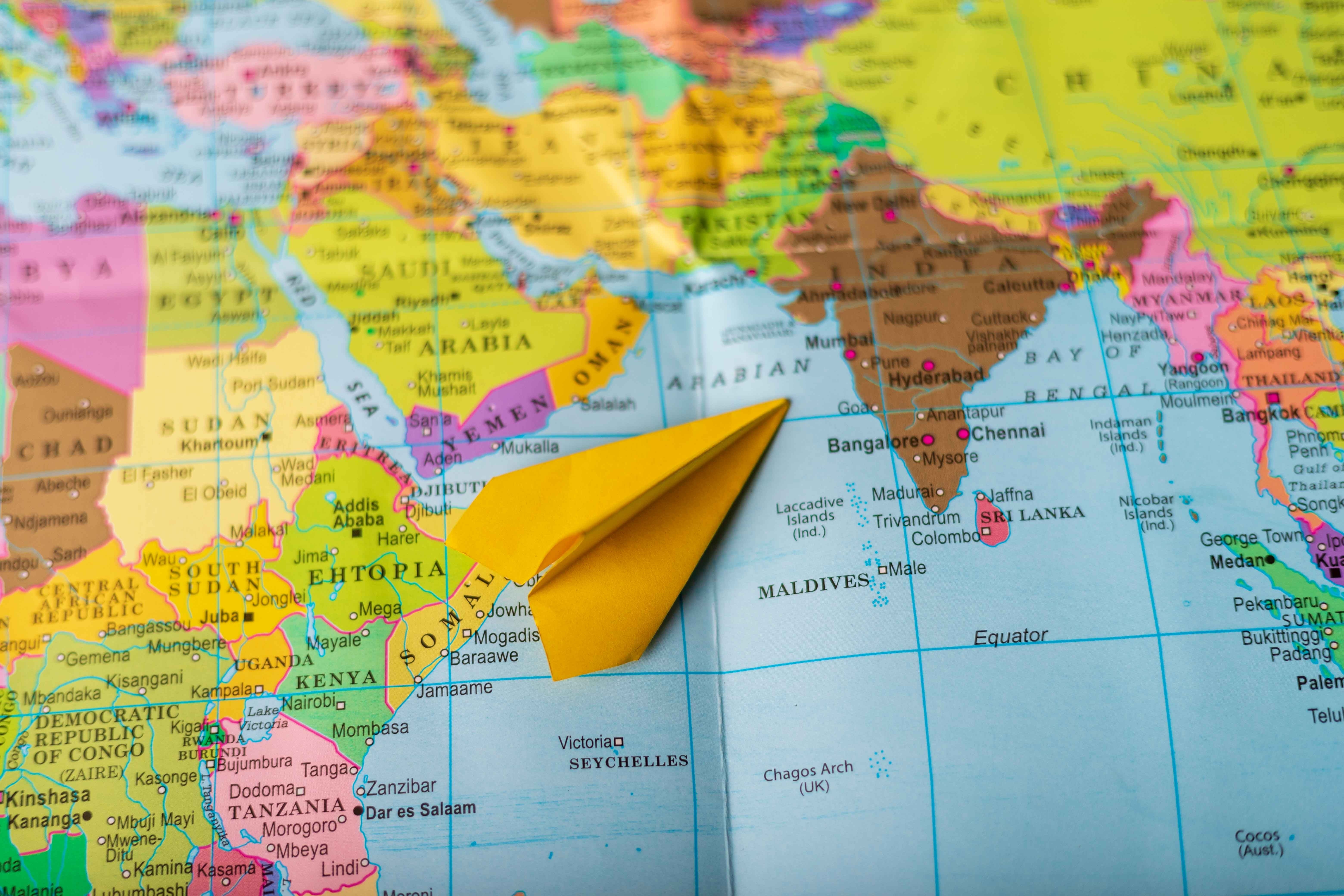 A yellow paper plane on a world map, positioned over the Indian Ocean near the Maldives and India, suggesting travel or exploration themes