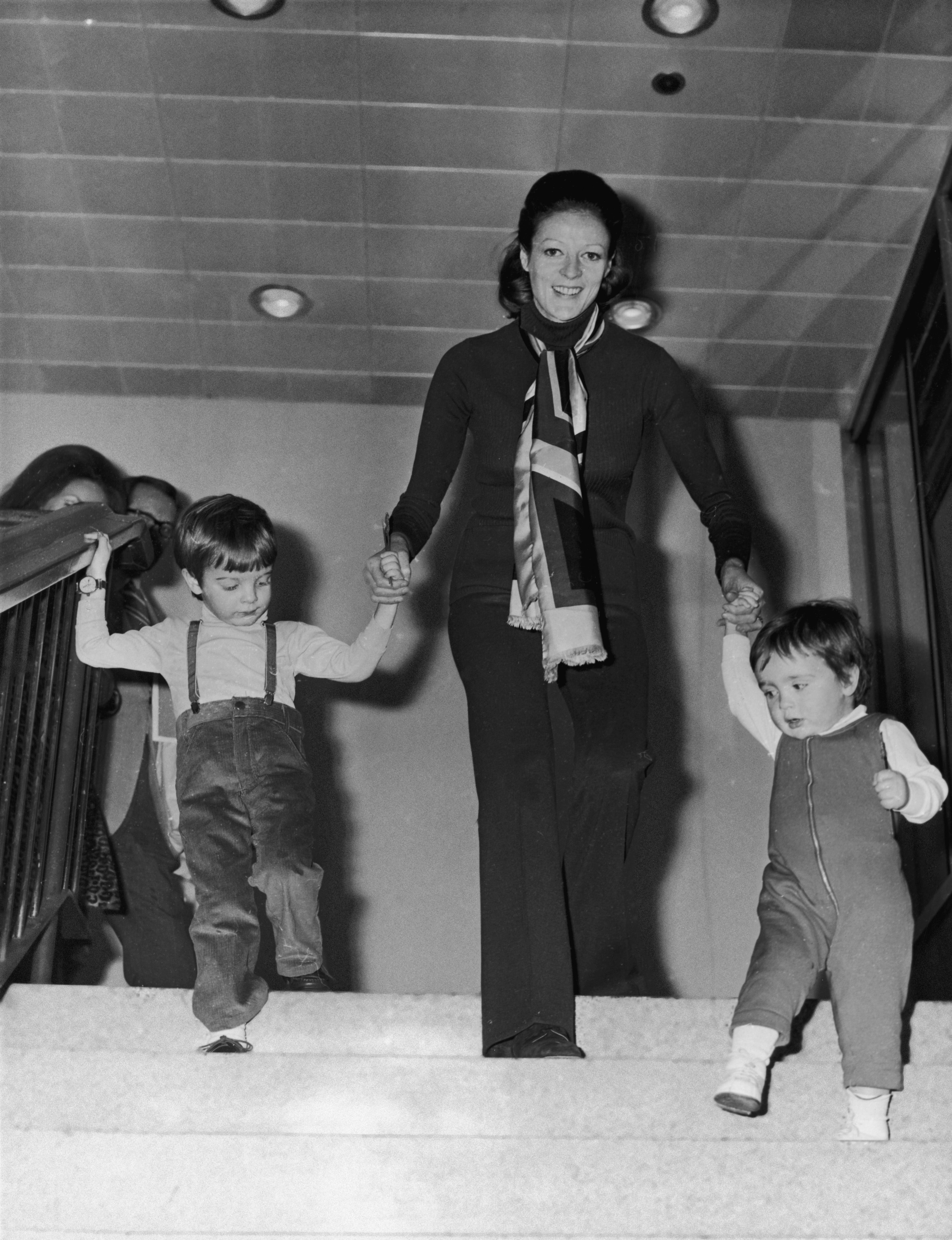 Maggie Smith guides 2 tiny children down stairs, holding each child's manus for support