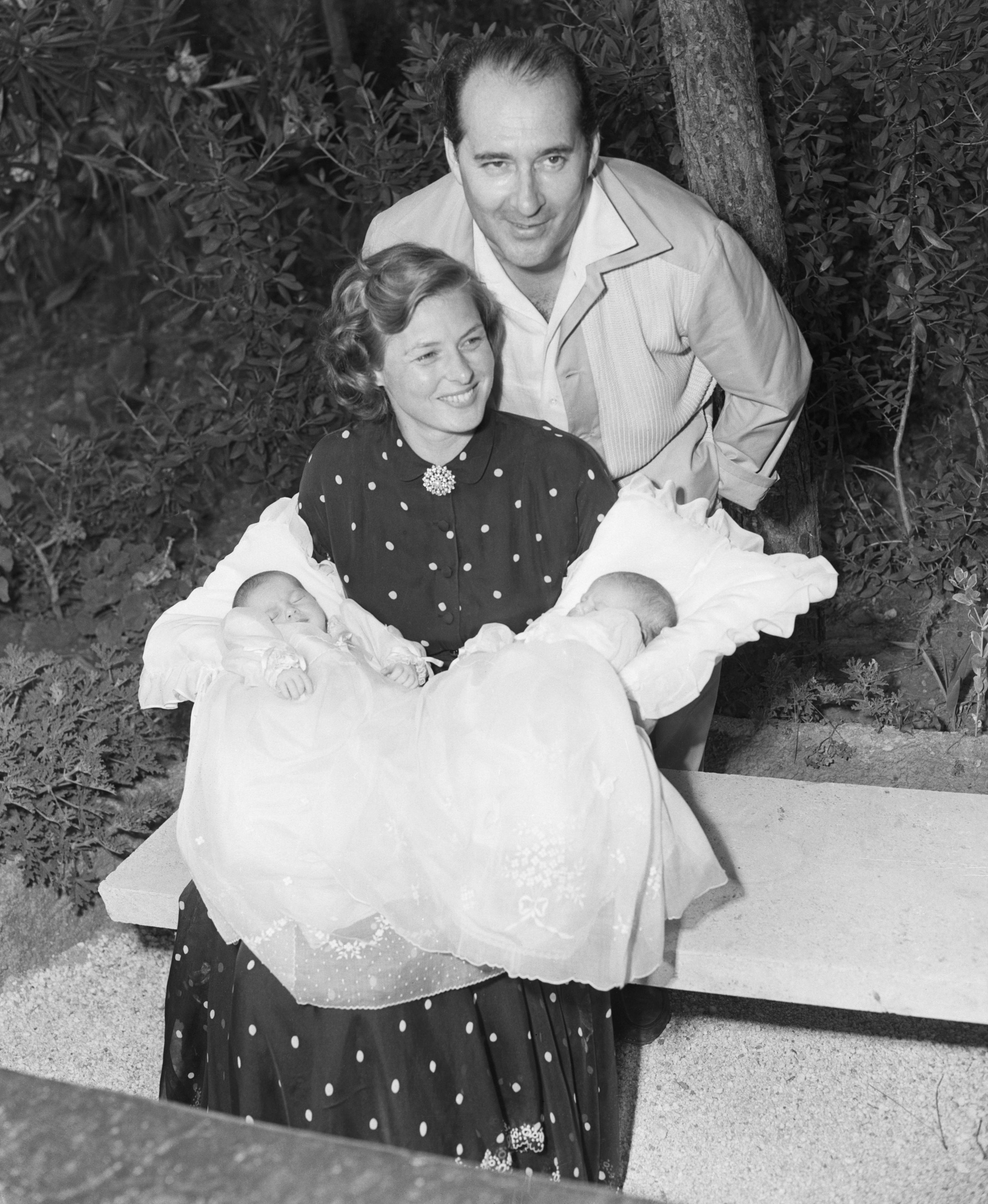 Ingrid Bergman and Roberto Rossellini grin astatine the camera, sitting connected a bench, Ingrid holding a babe successful each arm