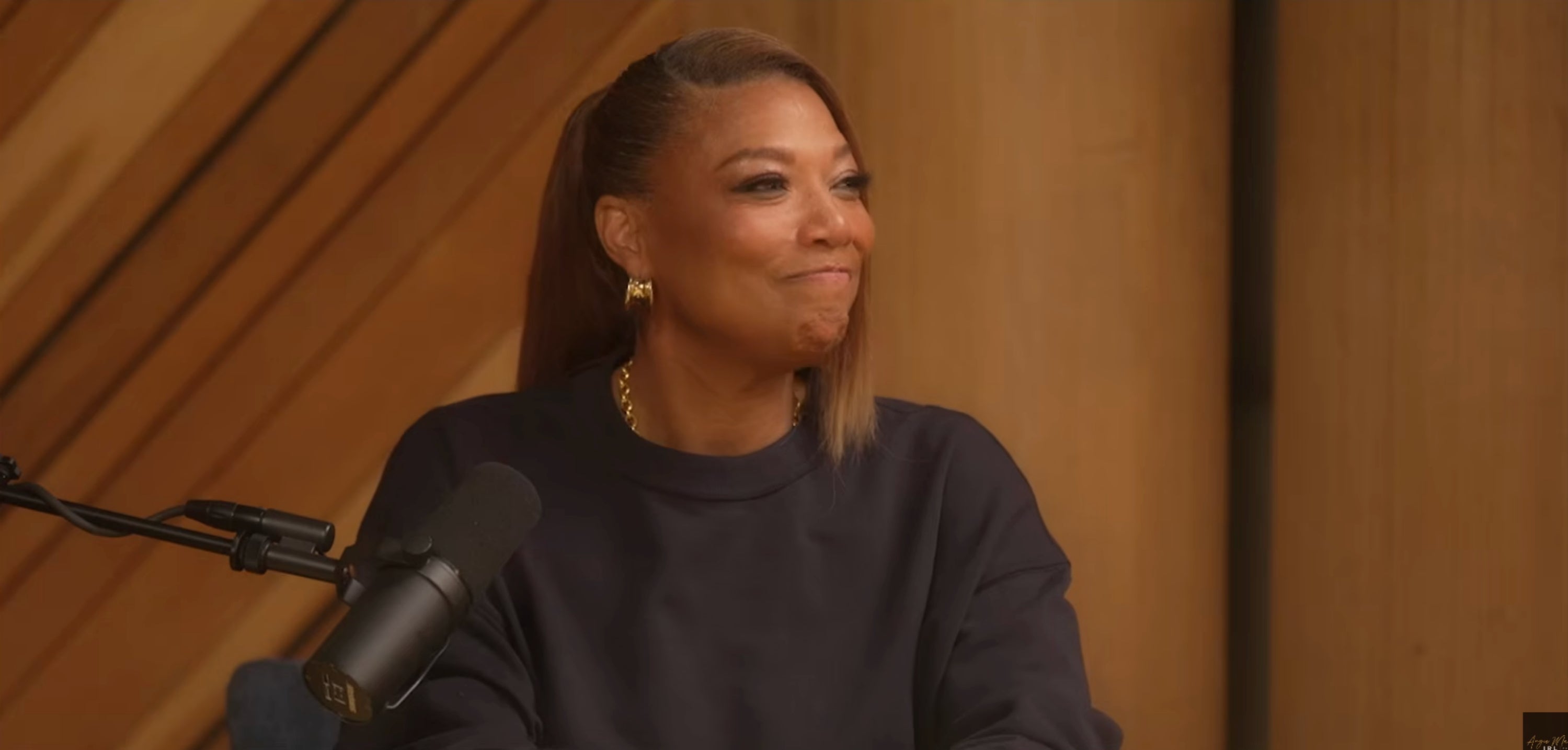 Person with a precocious ponytail and golden earrings, speaking into a microphone, wearing a casual sweater indoors