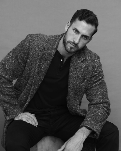 A man seated, wearing a tailored blazer over a casual shirt, looking directly at the camera with a relaxed pose
