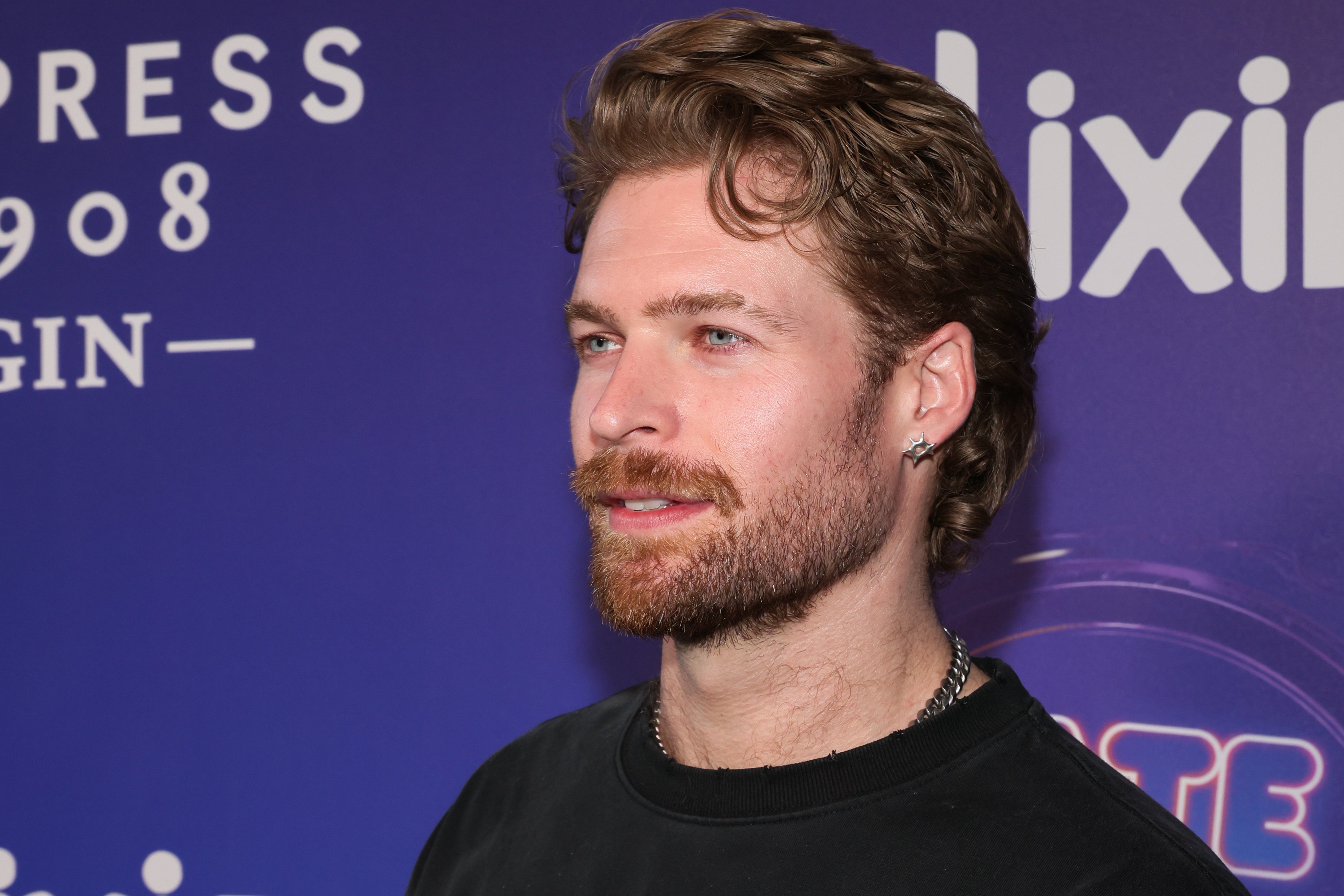 A person poses at a TV event, wearing a casual black top with styled hair and a beard, set against a logo backdrop