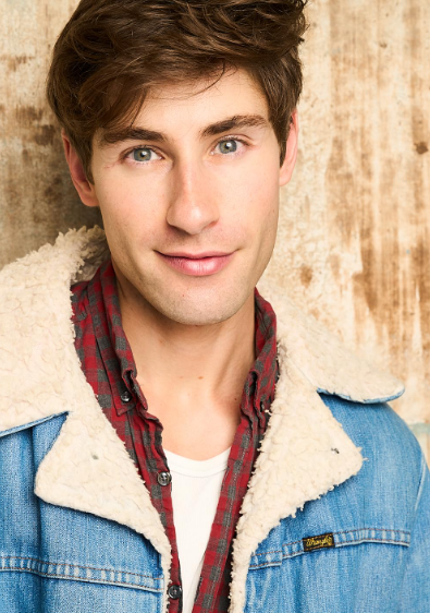 Person in casual attire with denim jacket and plaid shirt, smiling at camera. Neutral background suggests a relaxed setting