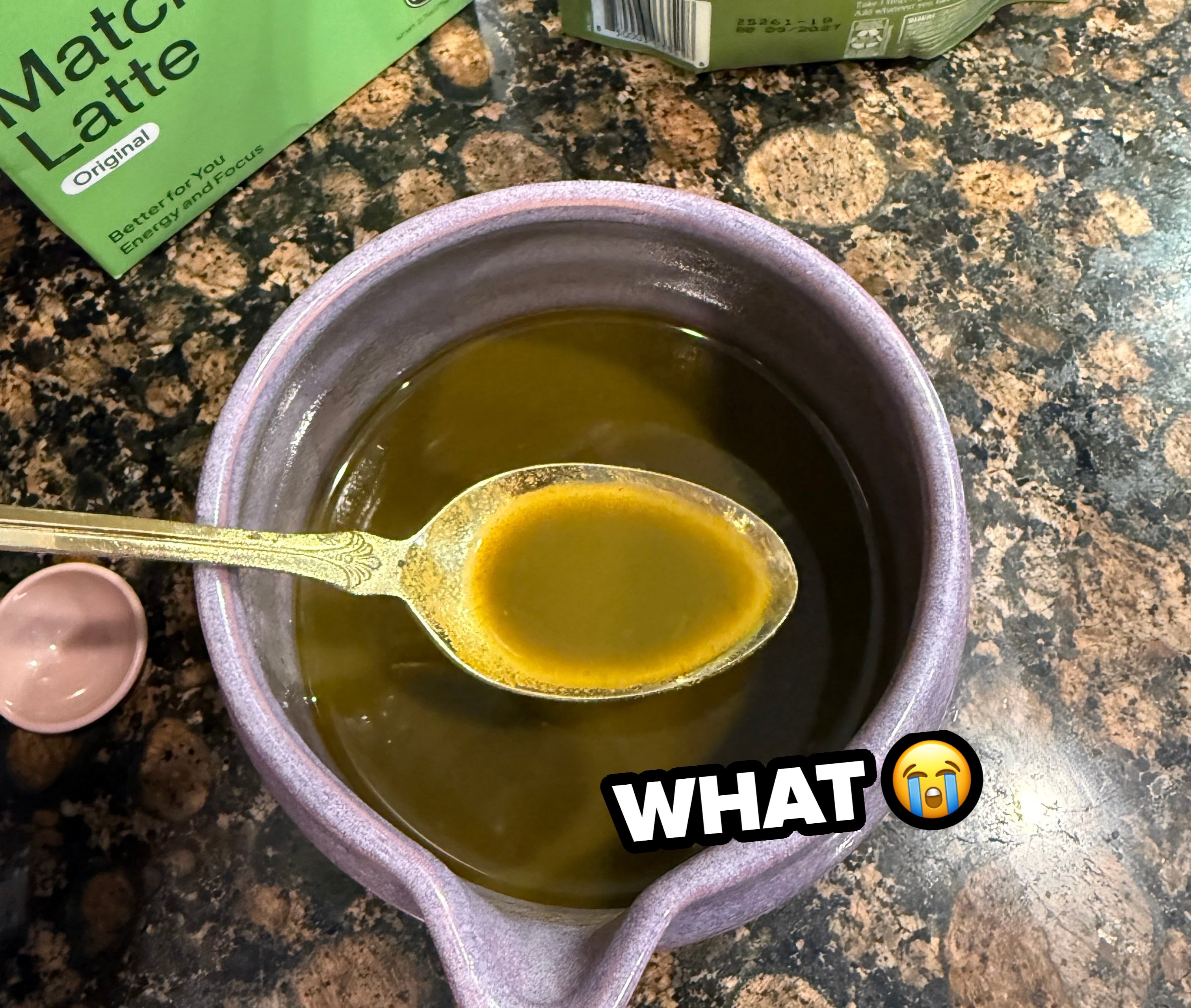 A purple bowl of matcha latte with a spoon. Matcha latte powder box is nearby on a stone countertop