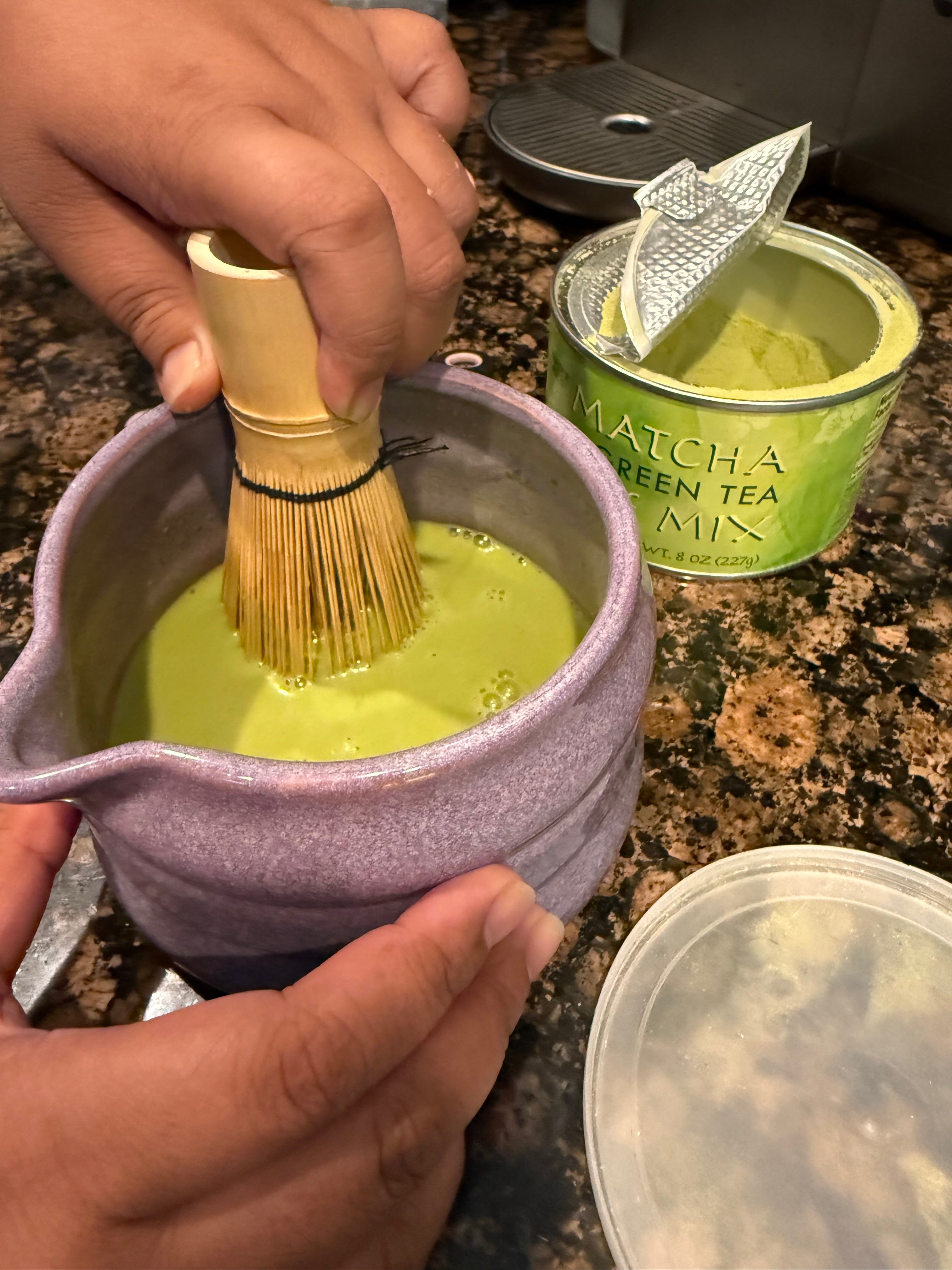 Hands whisking matcha green tea in a bowl next to an open matcha container on a countertop