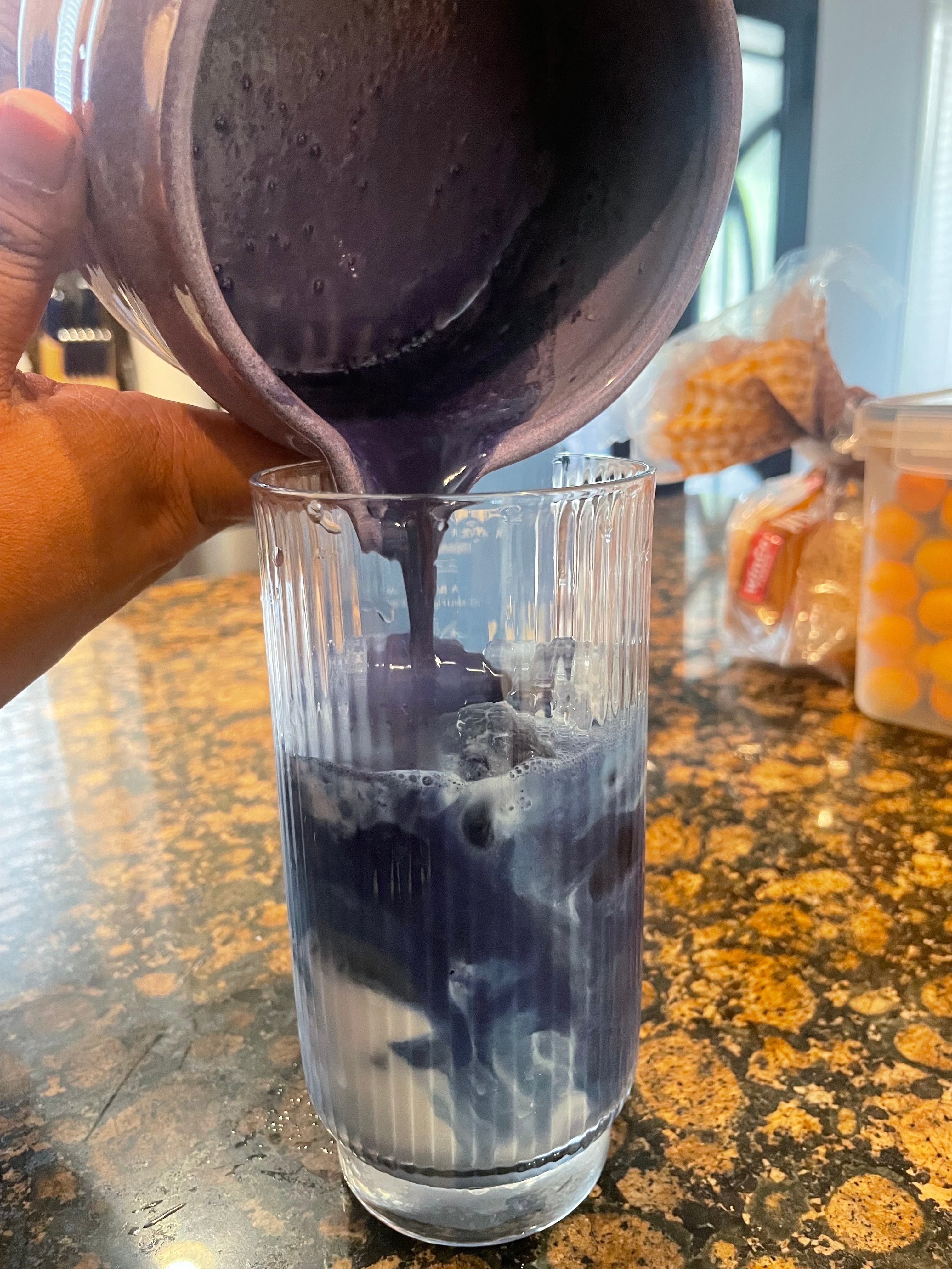 A person pours a thick purple smoothie into a tall glass on a kitchen counter